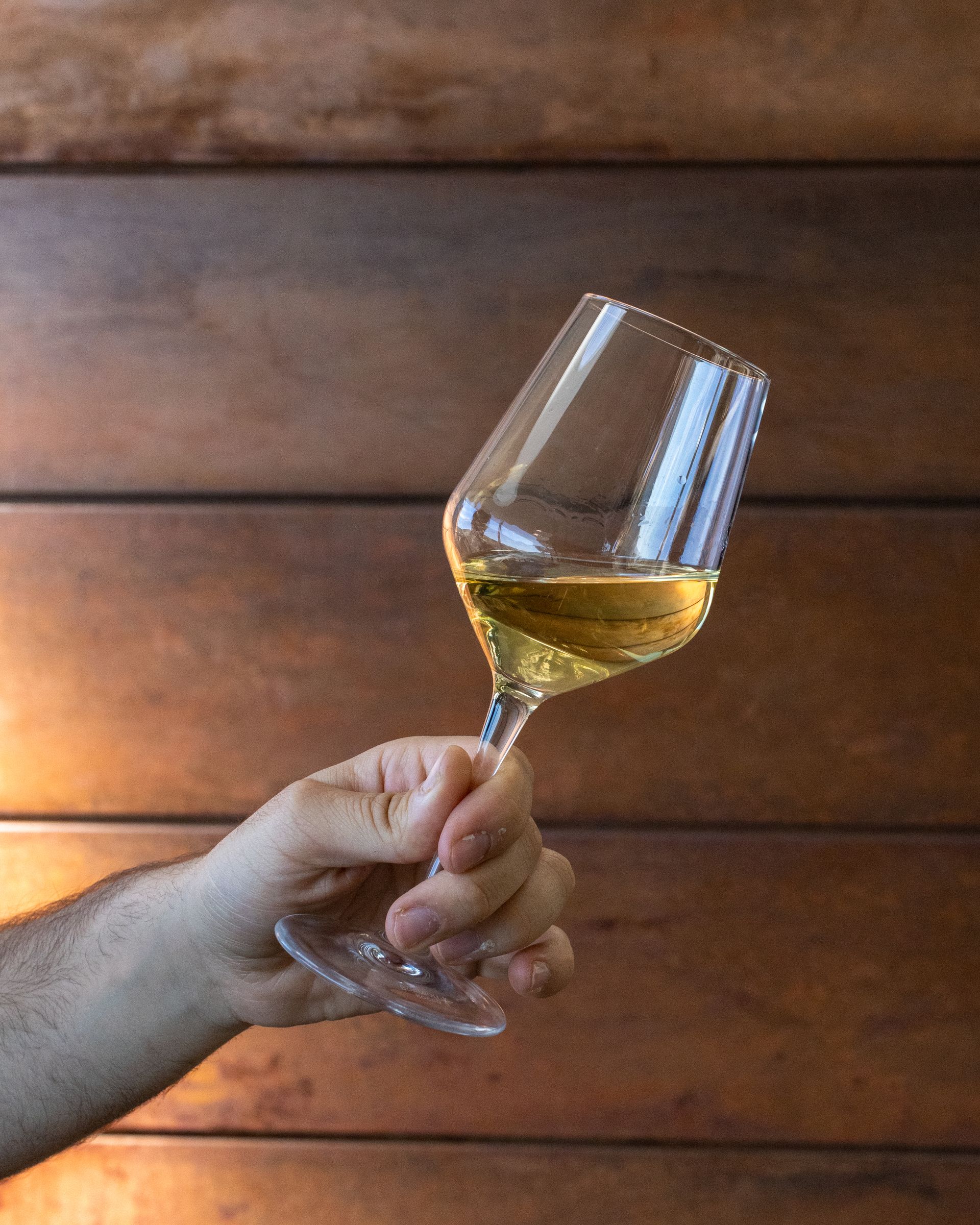 Una mano regge un bicchiere di vino bianco, appoggiato su uno sfondo di legno.