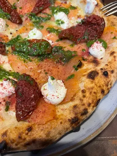 Primo piano della pizza: salmone affumicato, pesto, pomodori secchi, cucchiaiate di ricotta, su una crosta bruciacchiata.