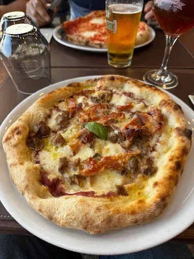 Pizza al piatto con salsiccia e formaggio, con un bicchiere di birra e vino sullo sfondo.