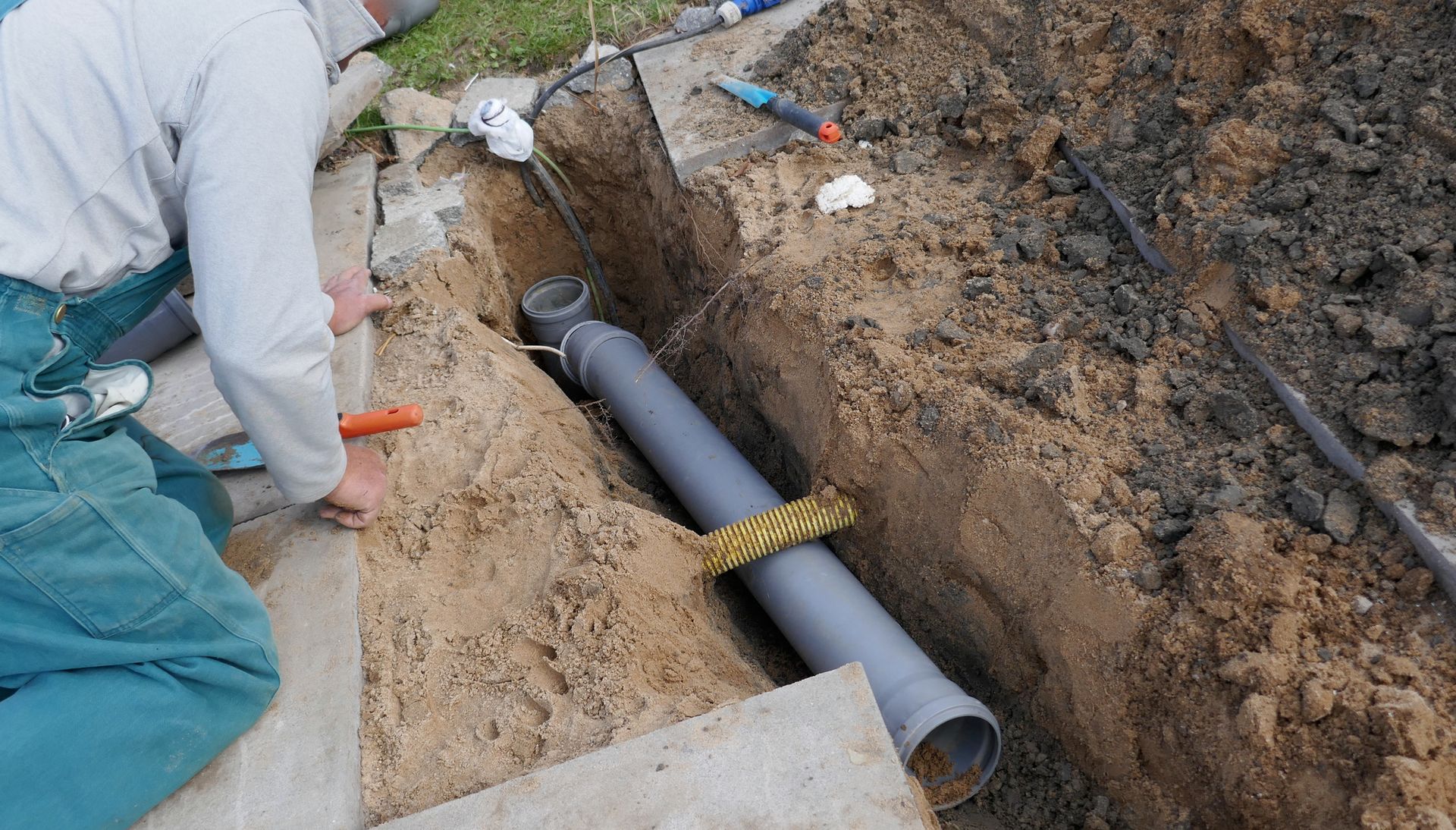 Person installing gray pipe in a trench; dirt and concrete nearby.