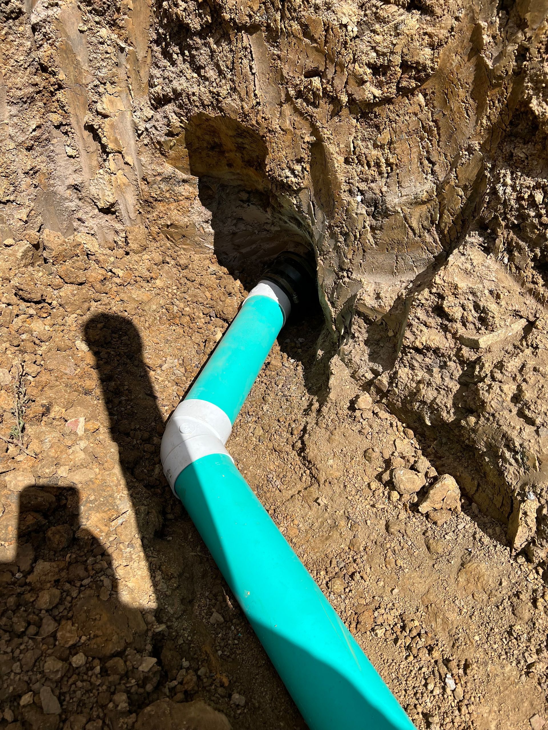 Turquoise PVC pipe with elbow fitting connected to a dark hole in a dirt wall.