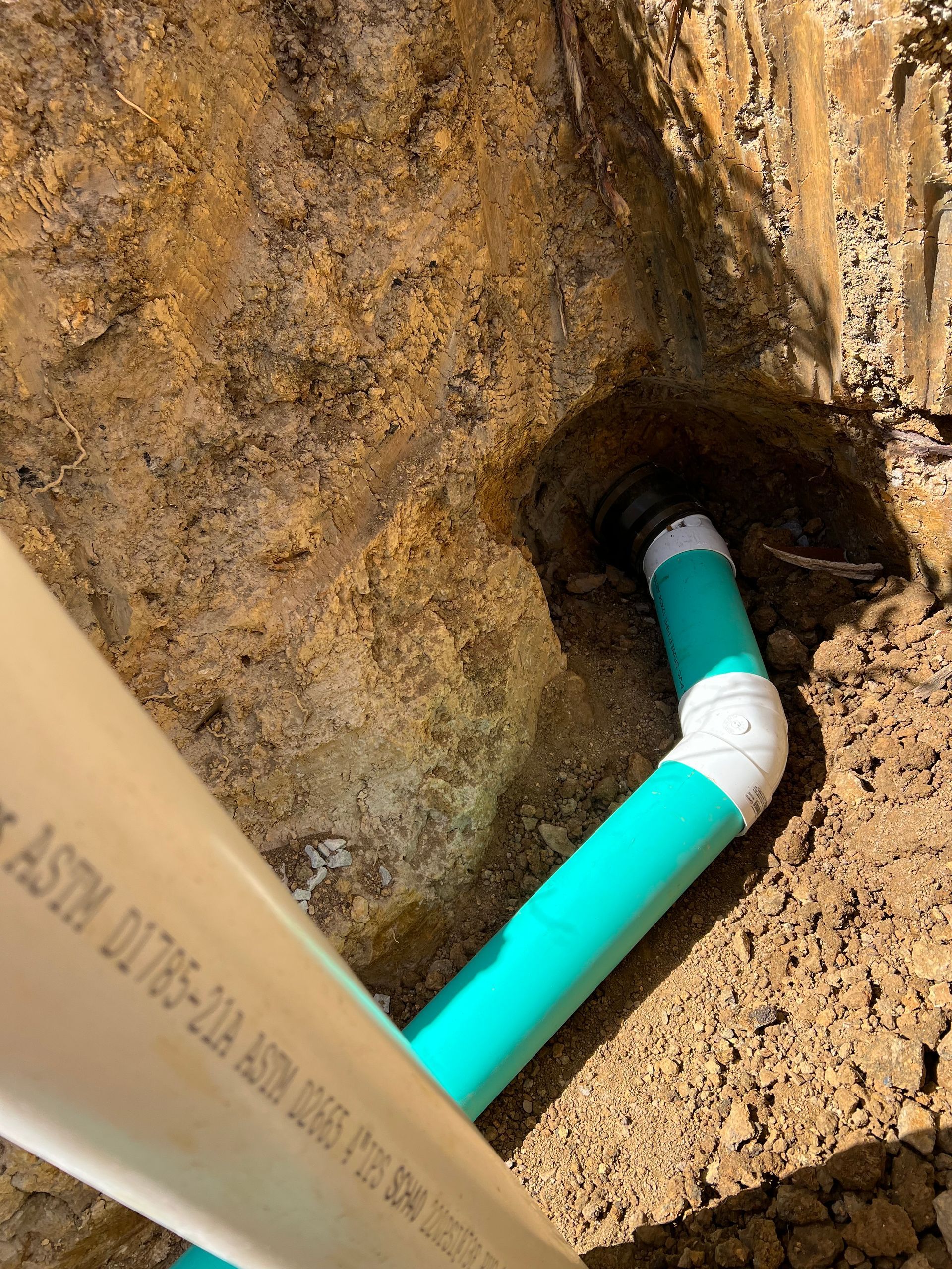 Green and white PVC pipes in a dirt trench, connected with a curved elbow fitting.