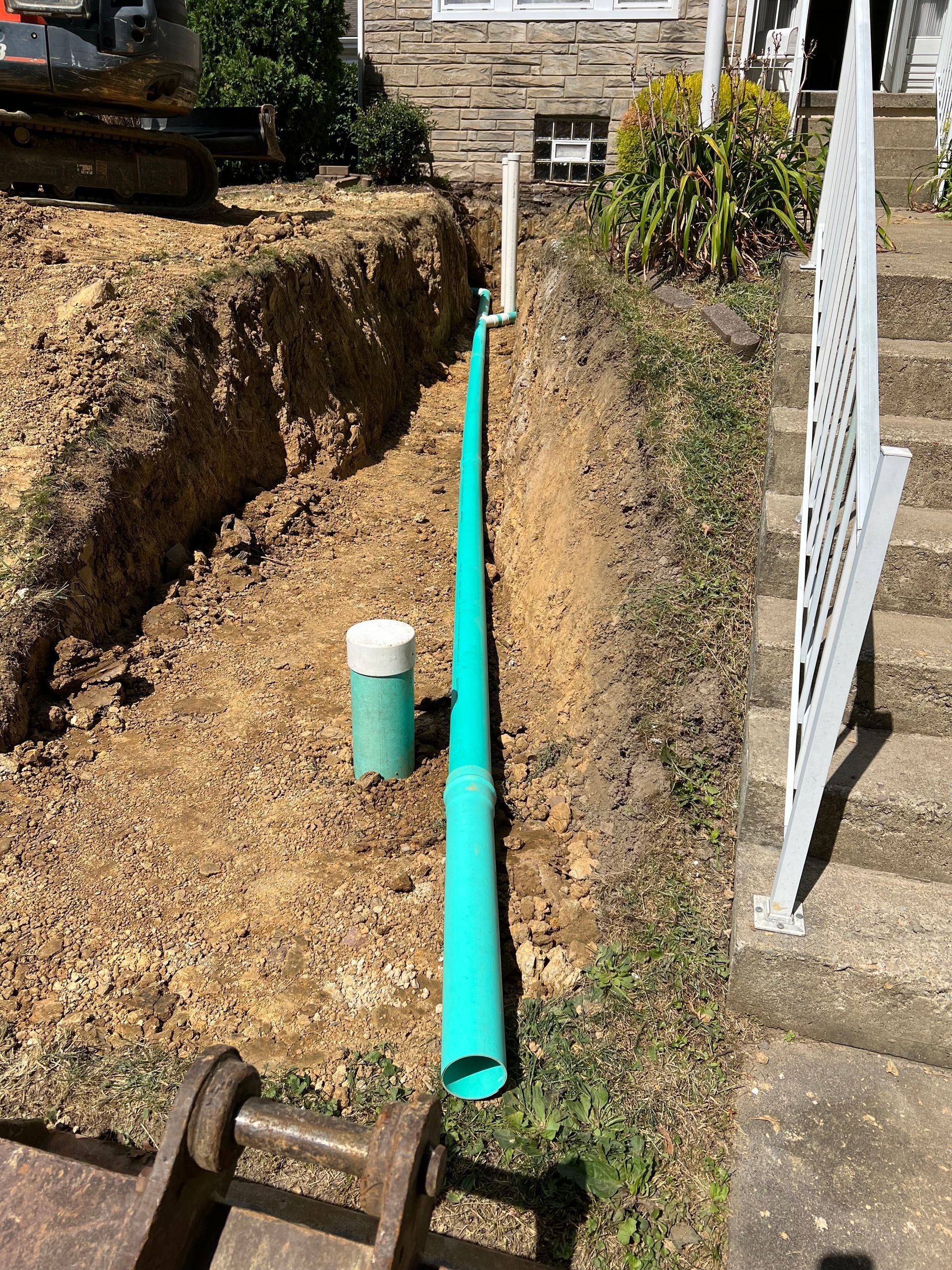 Ditch dug next to a house with exposed green piping, white cap, and a metal stair railing.