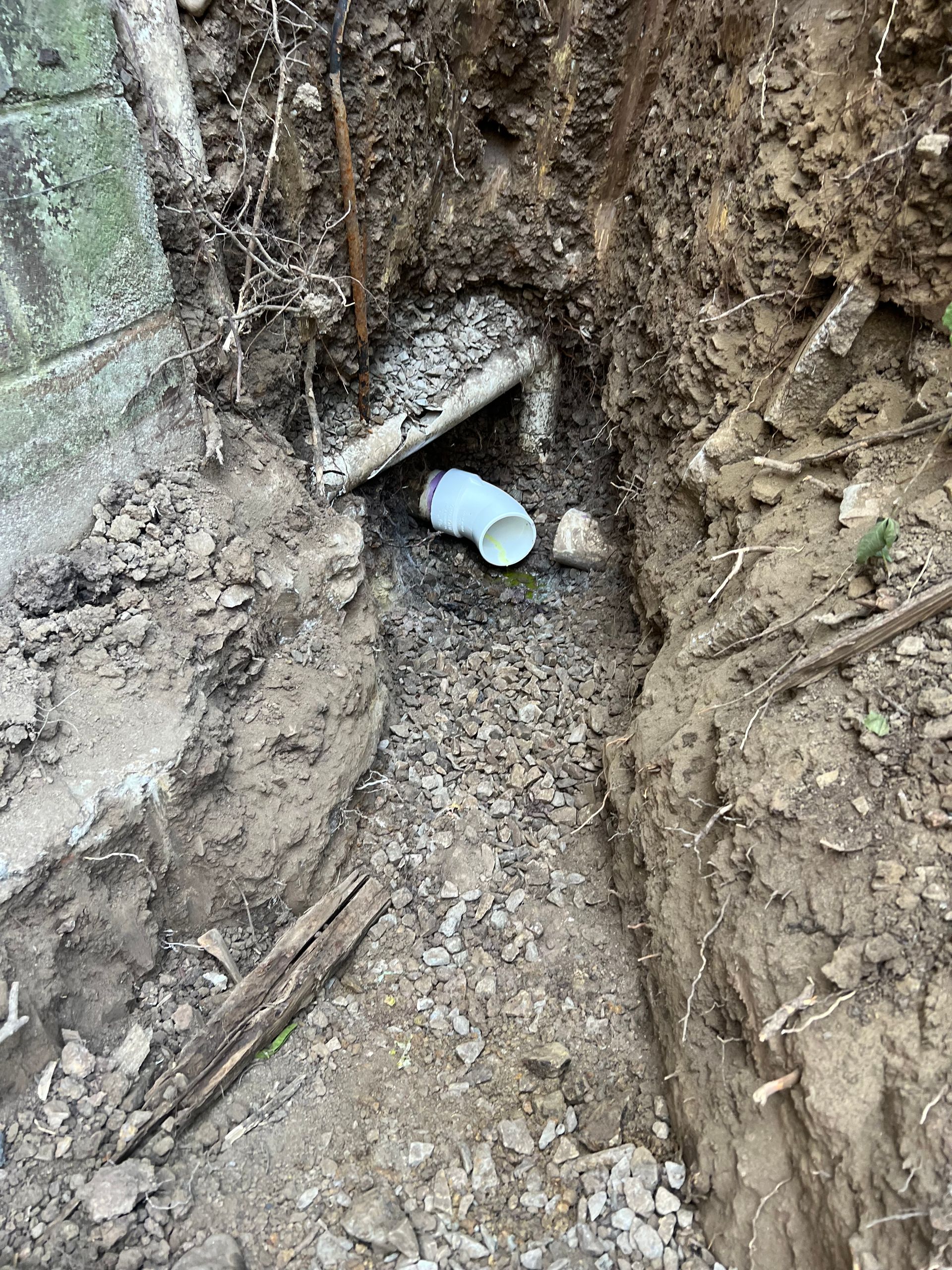 Trench dug near a concrete wall reveals a white PVC pipe. Dirt and gravel surround the pipe.