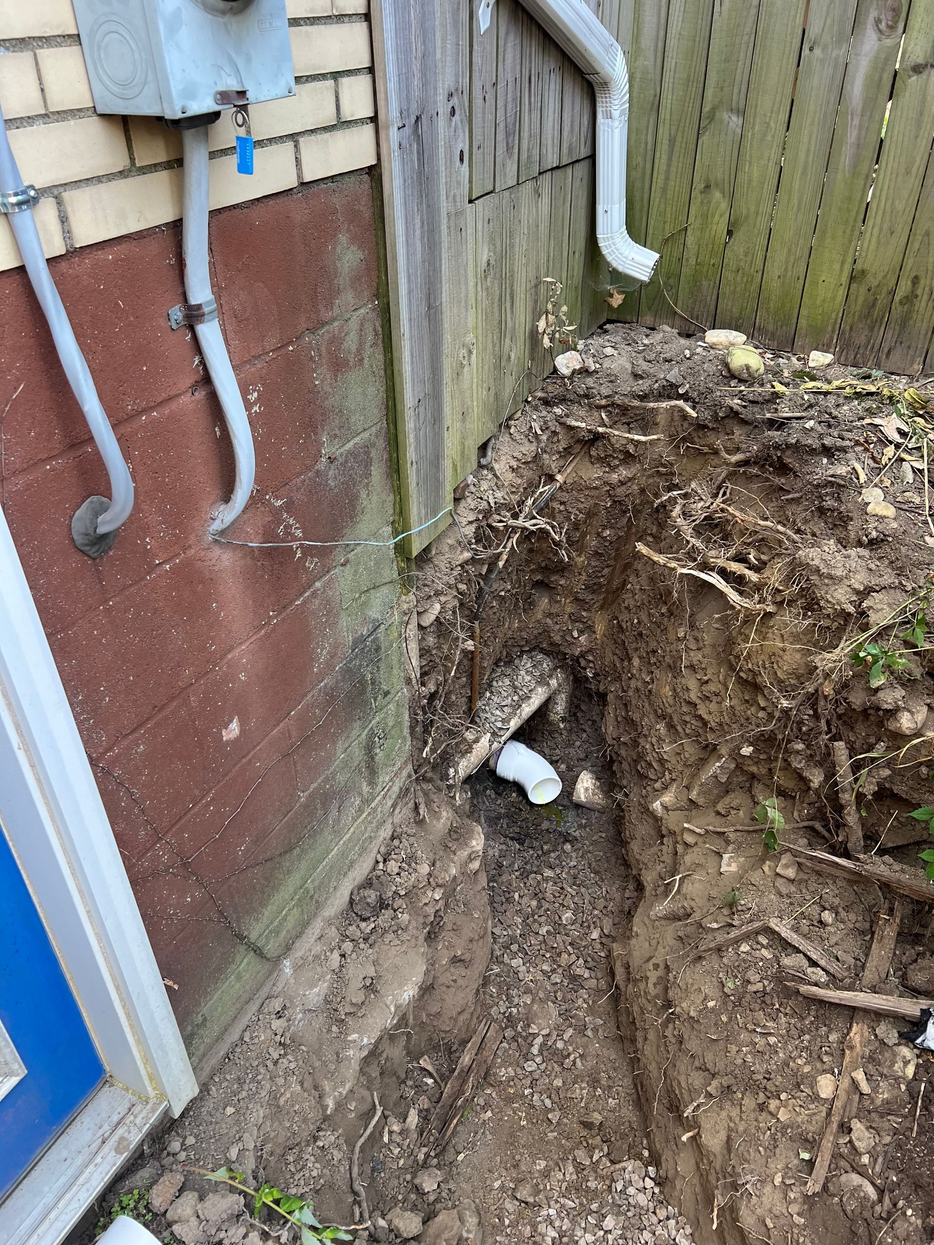Trench dug near a building and fence, revealing a white pipe. A gutter and electrical box are visible.