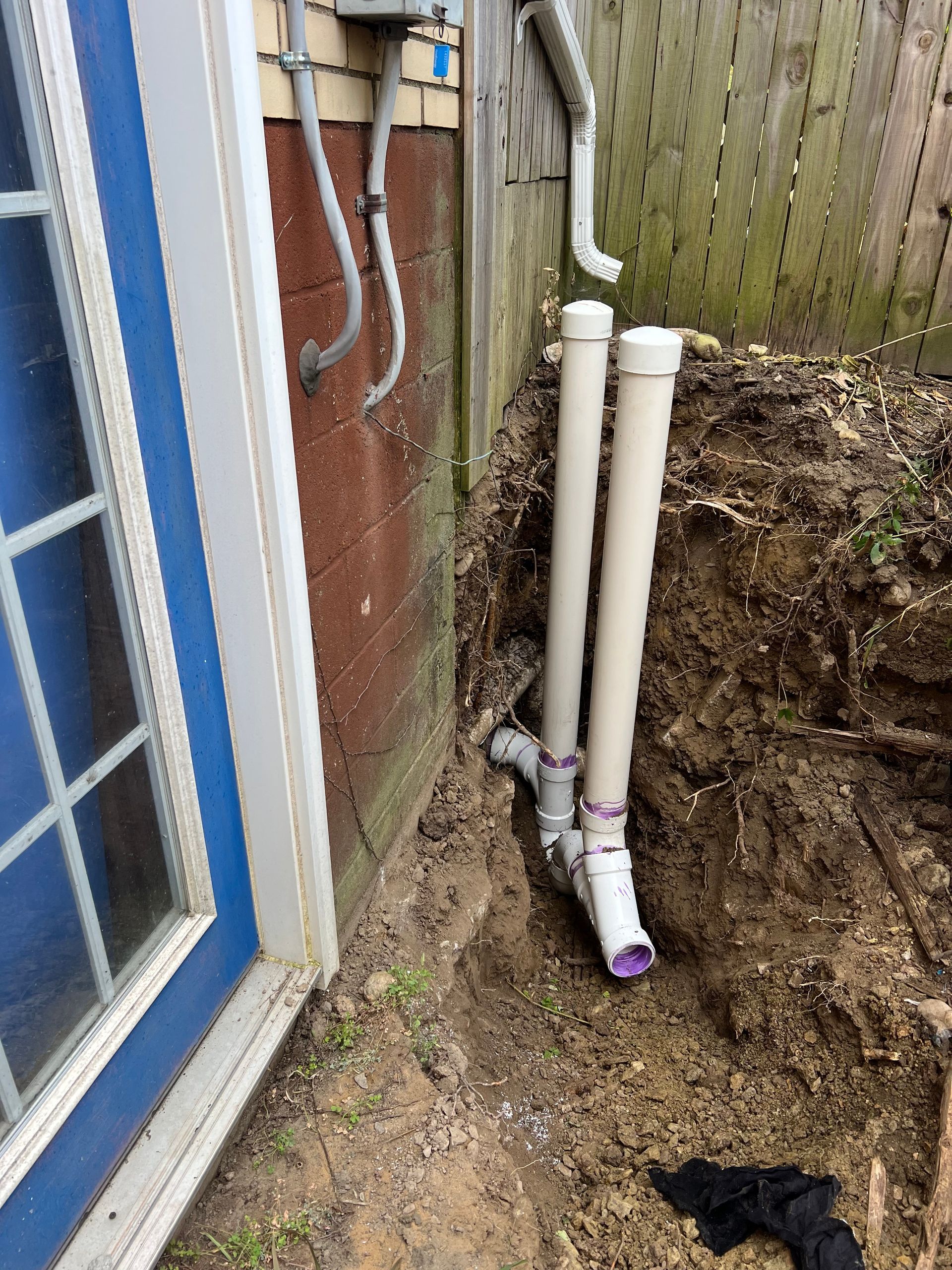 Plumbing pipes installed near a building, with a partially dug trench and a wooden fence in the background.
