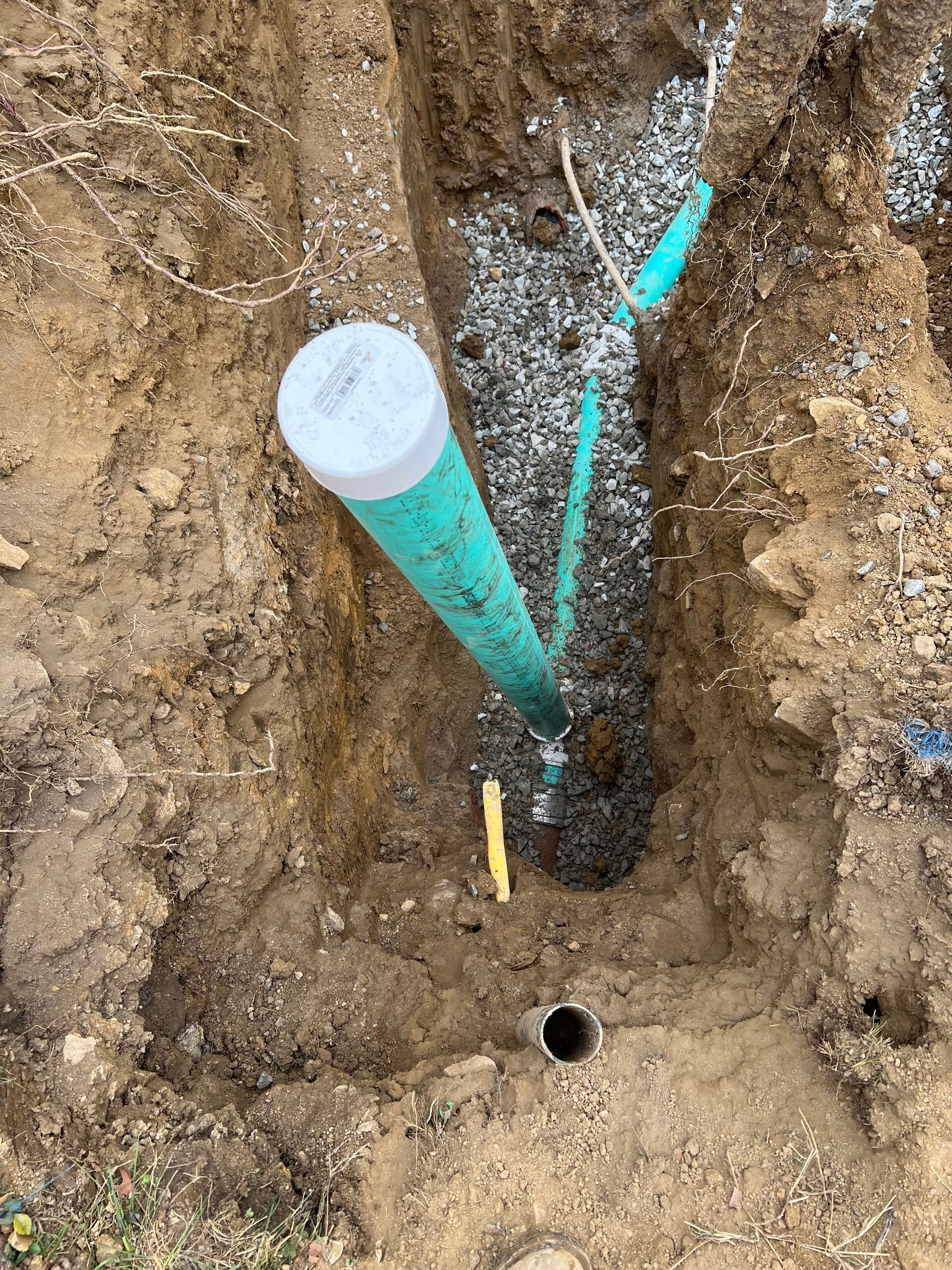Trench with green pipes and gravel. A large capped pipe stands vertically, leading to other buried pipes.
