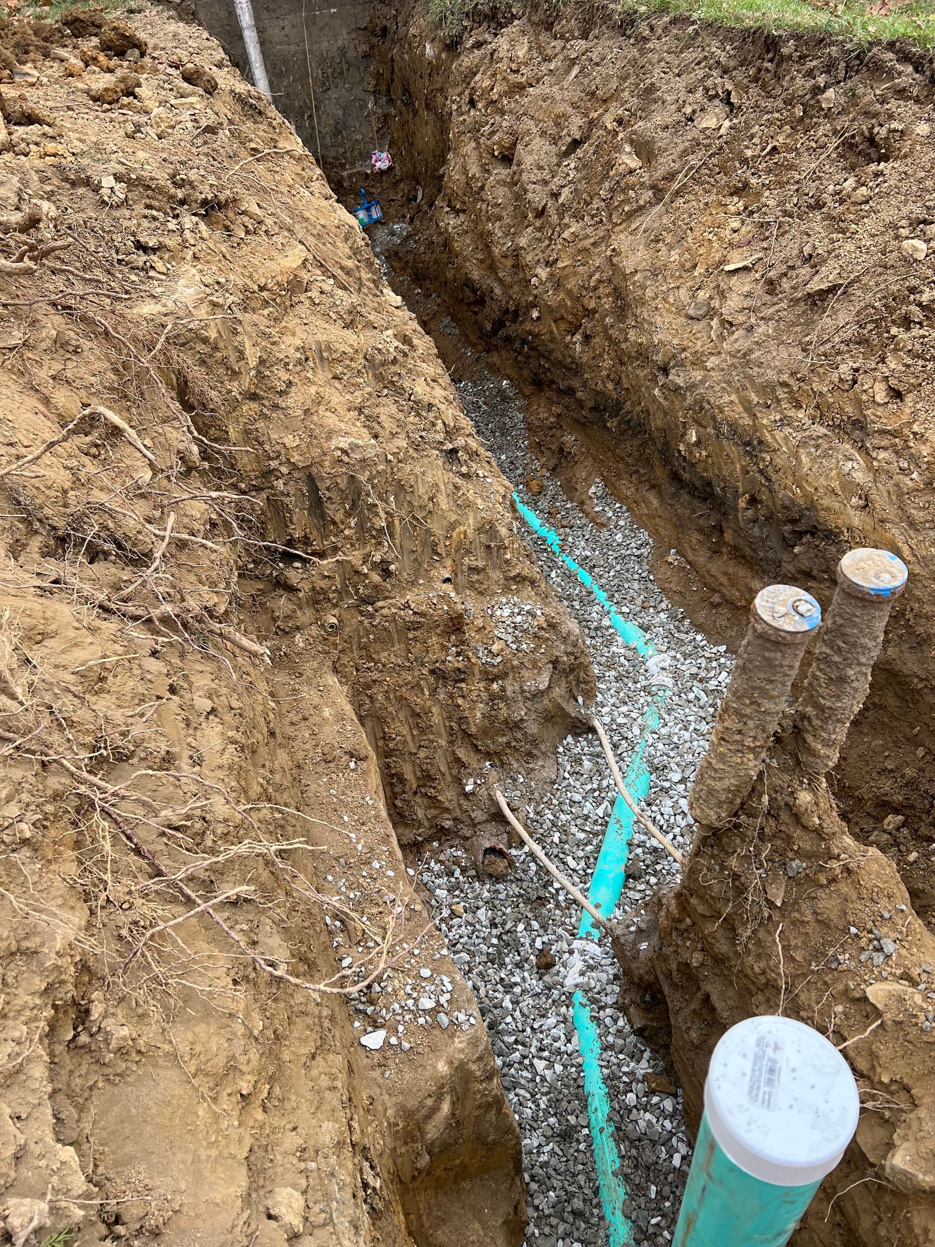 Trench filled with gravel, holding turquoise pipes. Two upright PVC pipes visible, soil walls.