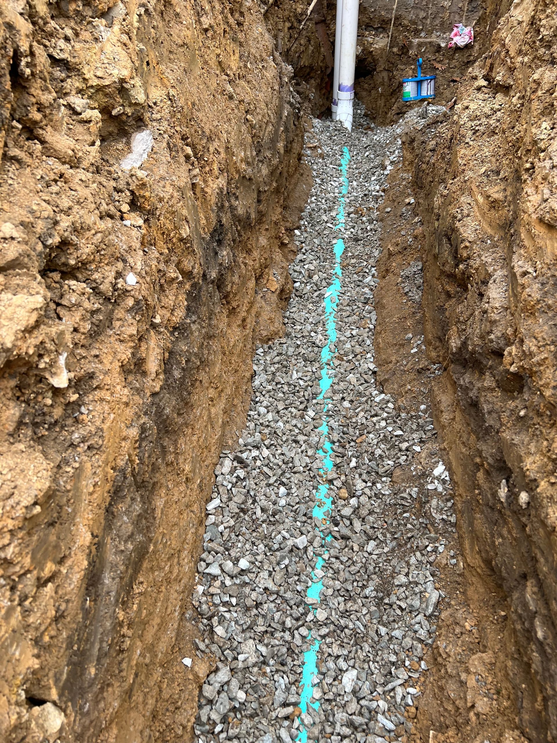 A trench filled with gravel, a blue pipe running through it, and a white pipe at the top.