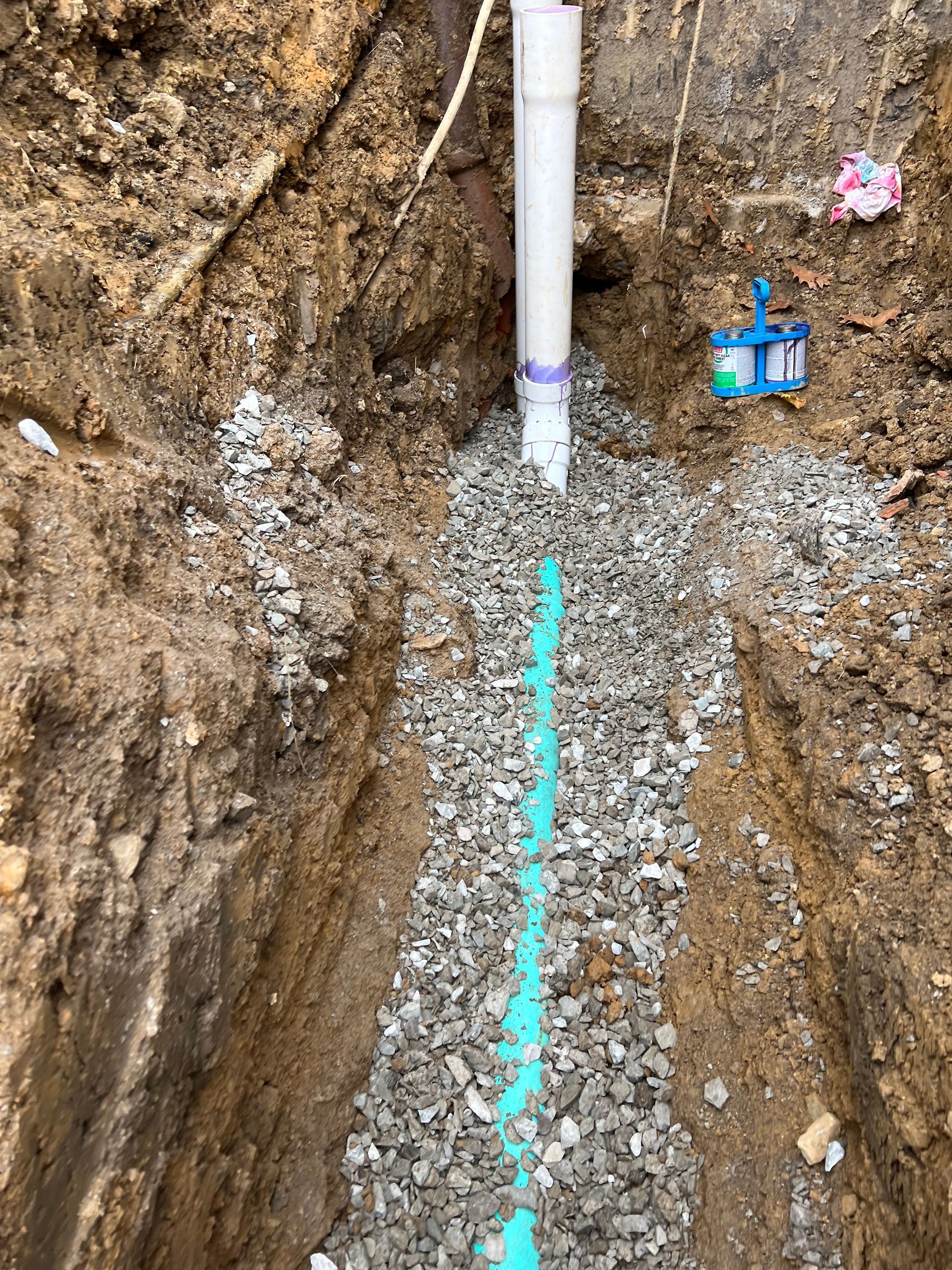 Trench with gravel and blue pipe, white pipe at top, likely drainage or utility installation.