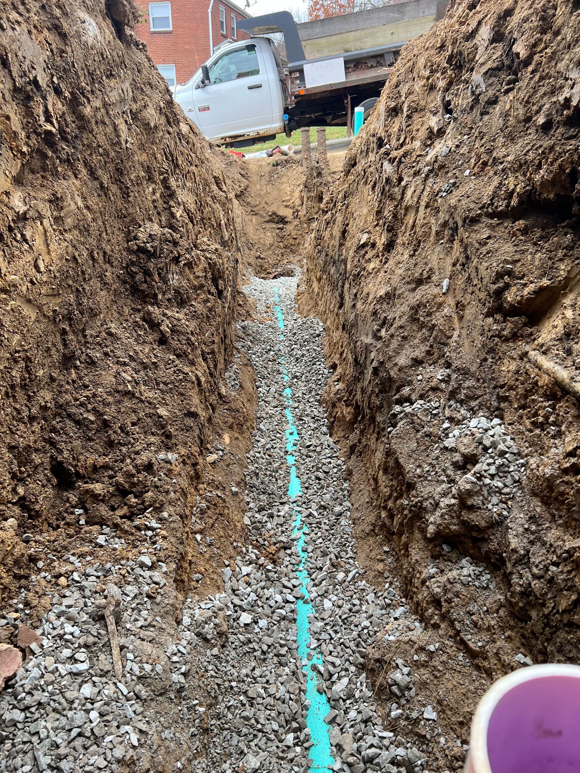 A trench with a blue pipe and gravel, likely for drainage, dug in the ground near a white truck.
