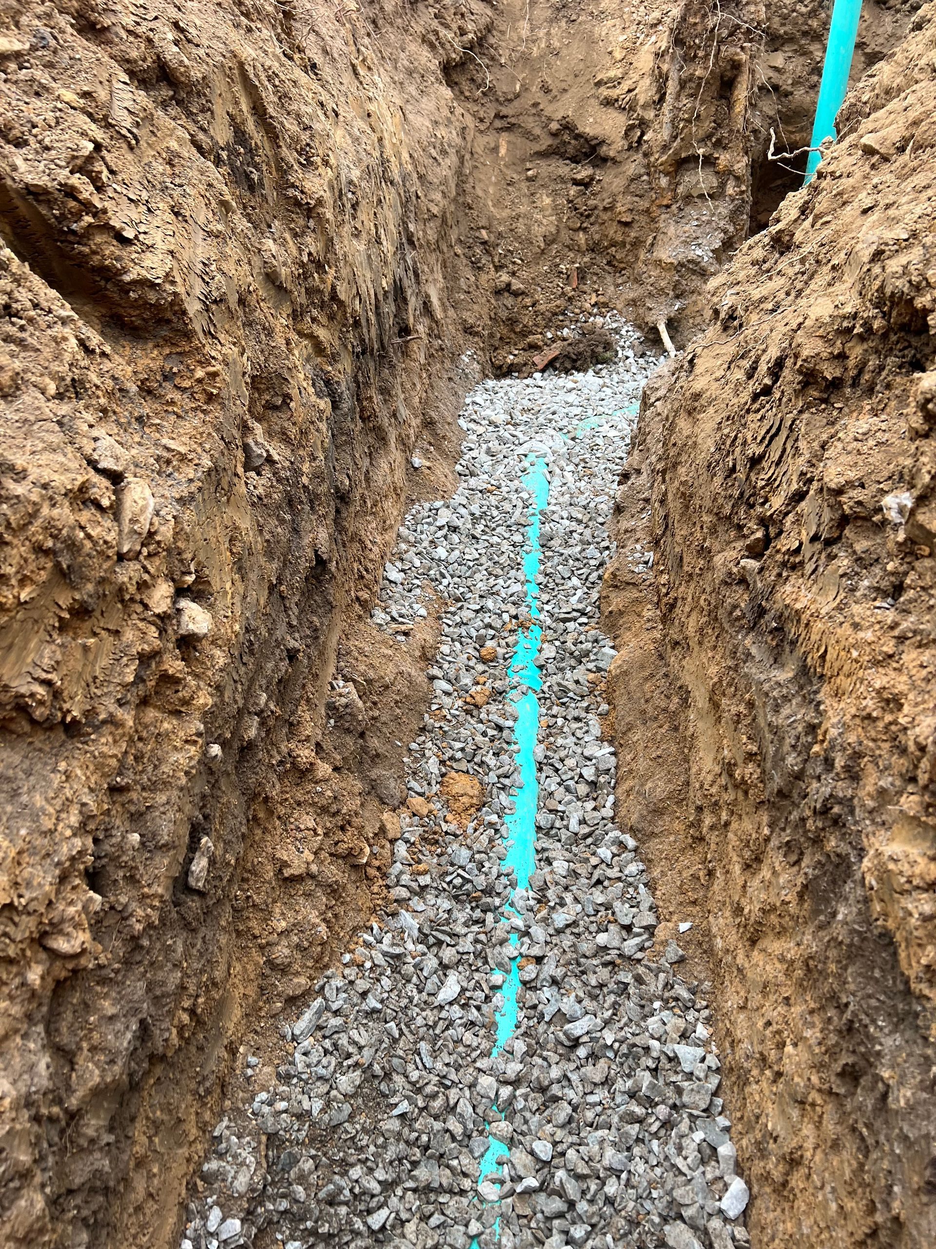 Trench with blue pipe and gravel fill, ready for burial; brown soil walls.