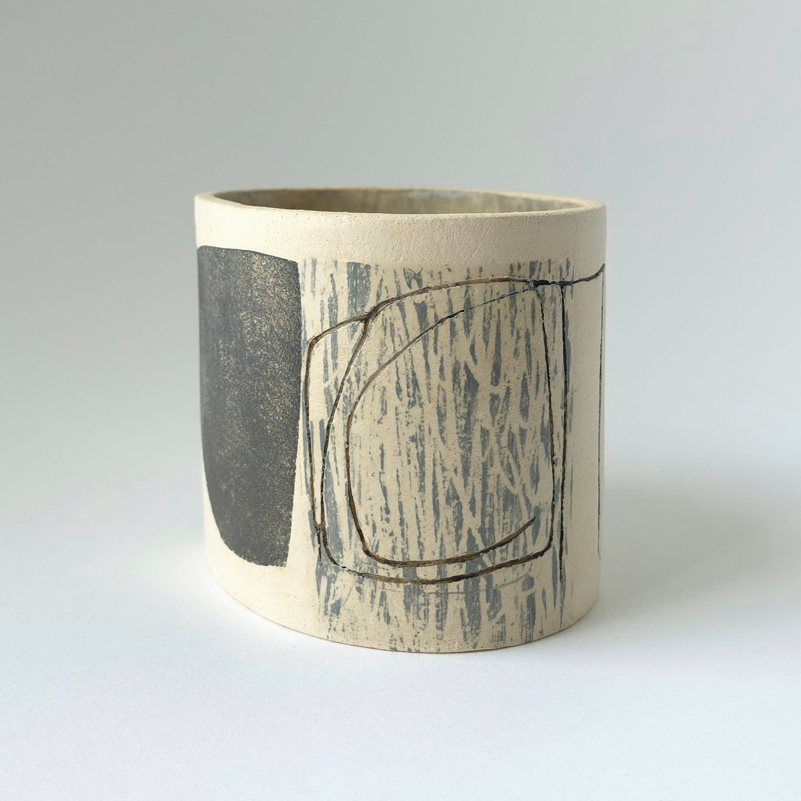 A cylindrical ceramic cup with a light beige speckled surface, featuring a solid dark grey shape and abstract etchings.