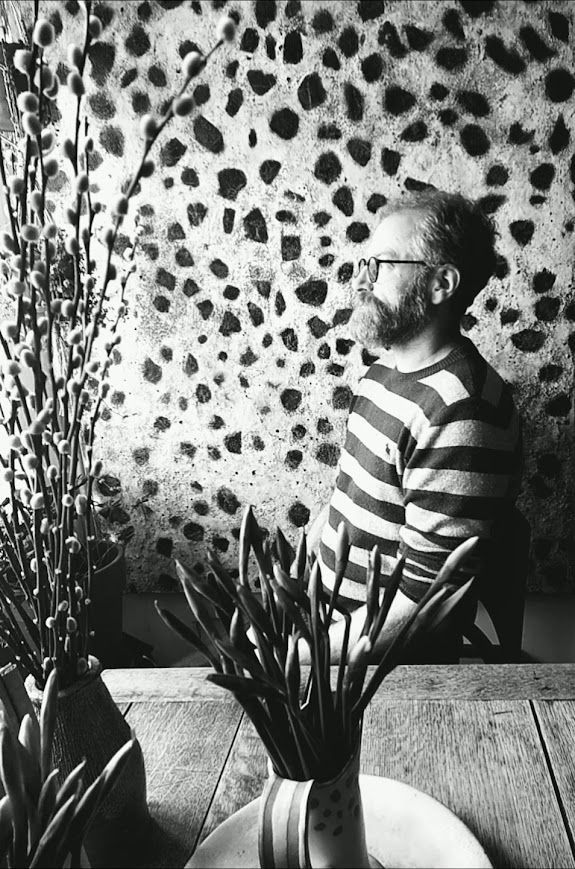 A man with a beard is standing in front of two paintings