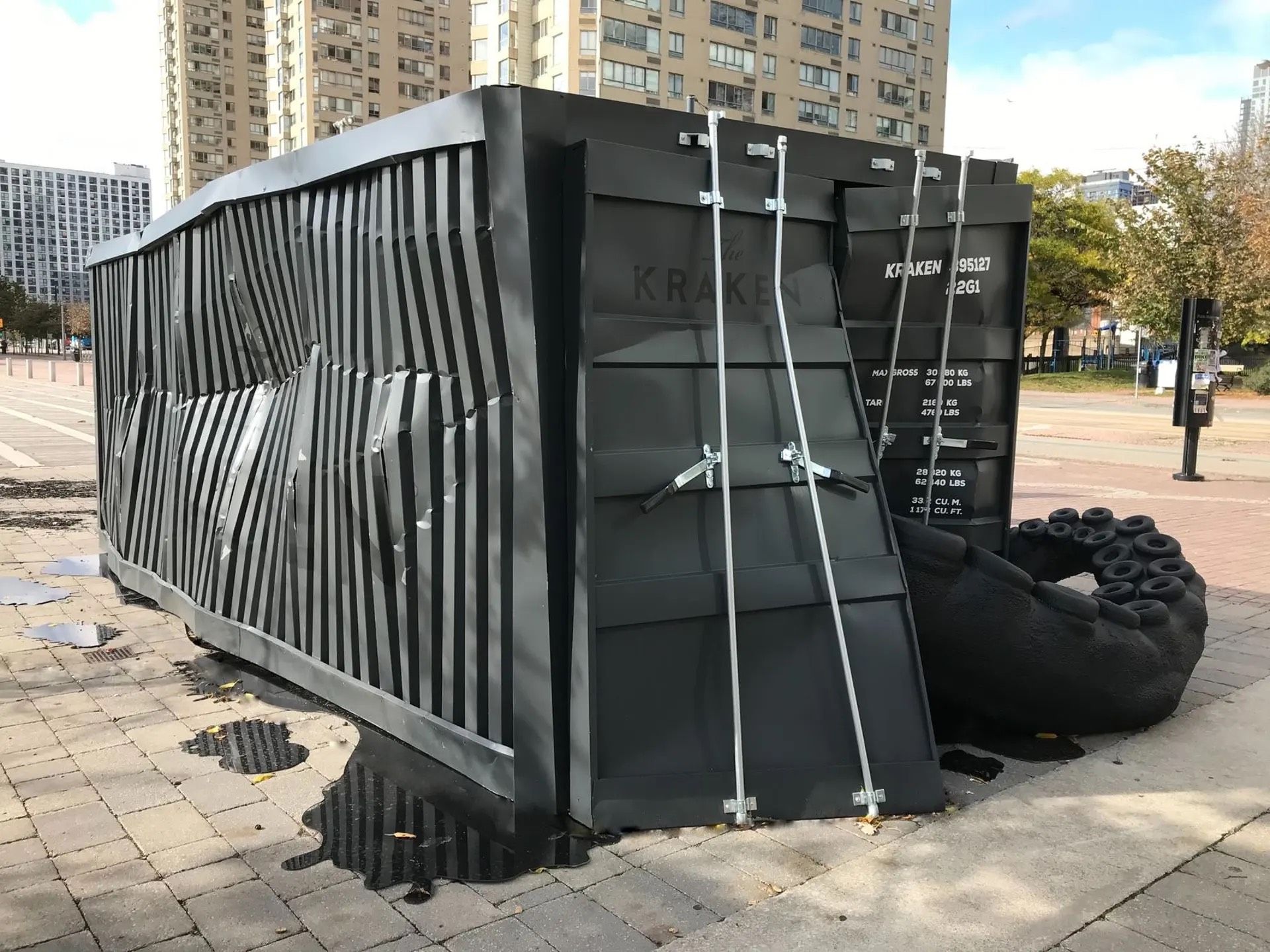 A black shipping container is sitting on the sidewalk next to a sculpture.
