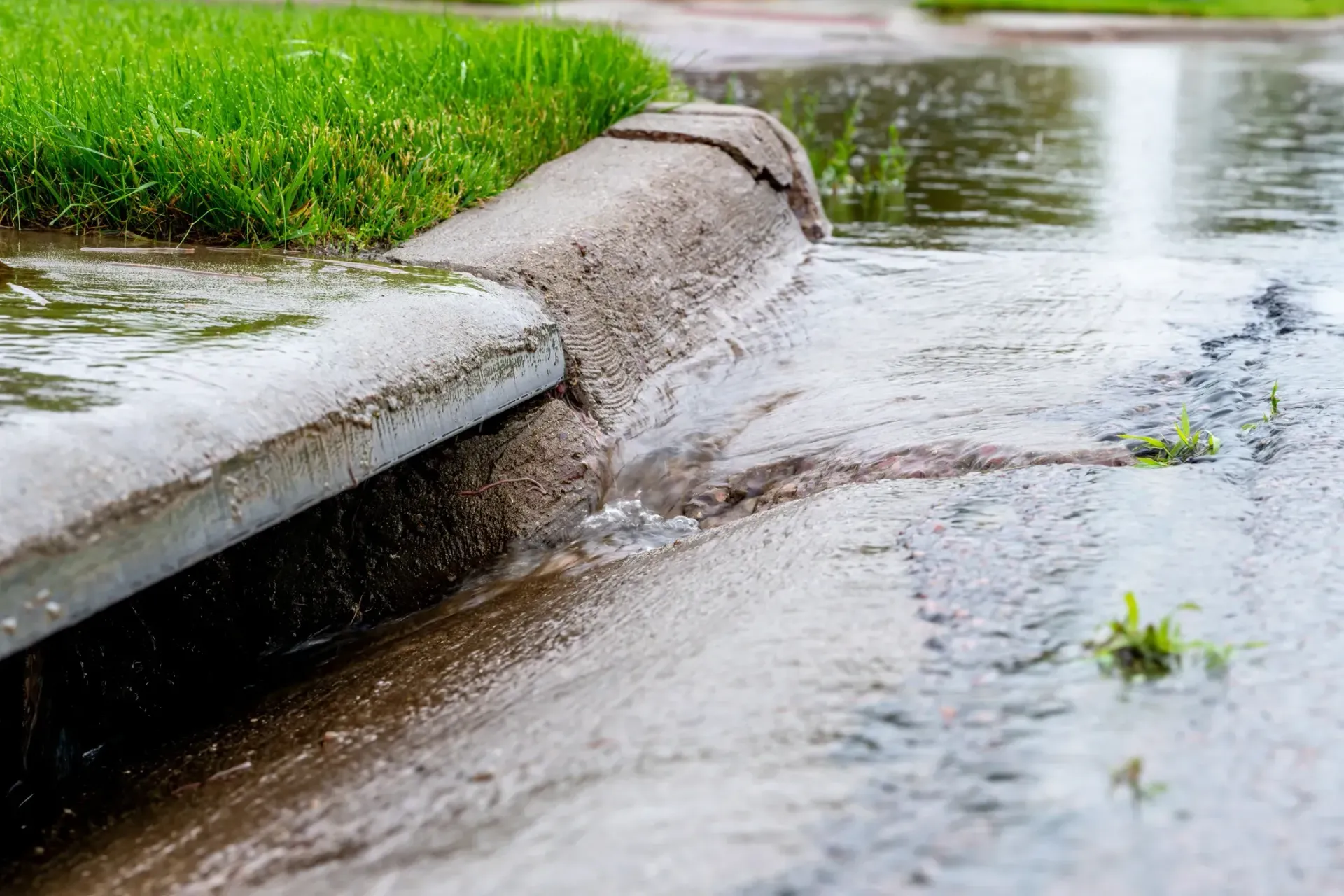 Rainwater flows from a green lawn over a cracked concrete curb into a flooded street.
