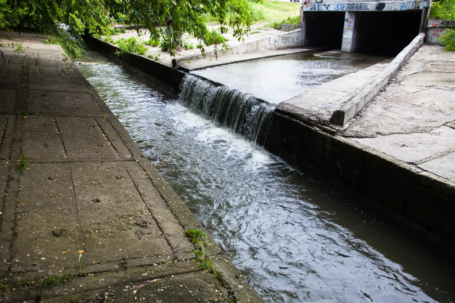 Water flows over a small concrete weir into a channel leading toward two dark tunnel entrances under a road.