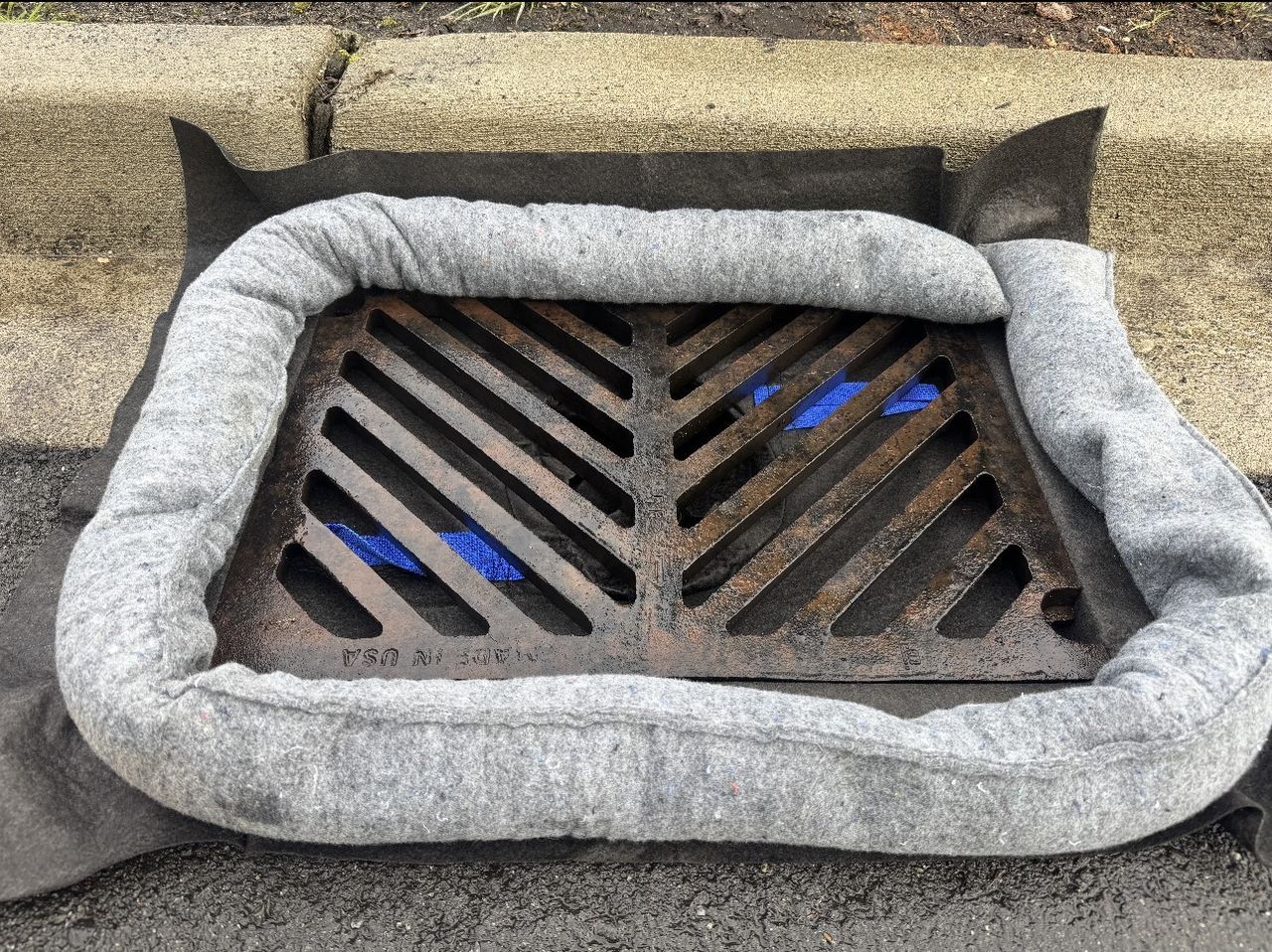 A storm drain grate covered by a grey, felt sediment filter with a protective absorbent boom around its edges.