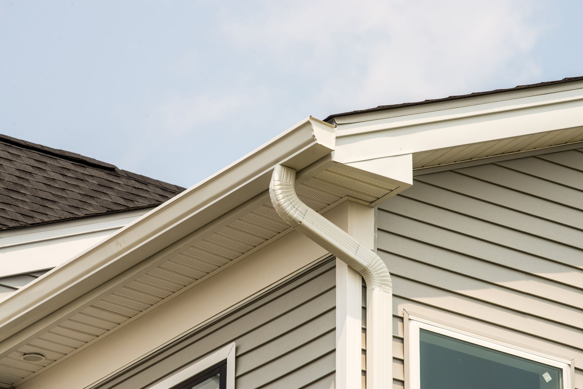 Des gouttières blanches et une descente pluviale fixées à l'angle d'une maison au revêtement beige et au toit en bardeaux.