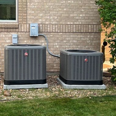 Two gray outdoor air conditioning units beside a brick house wall on a grassy lawn