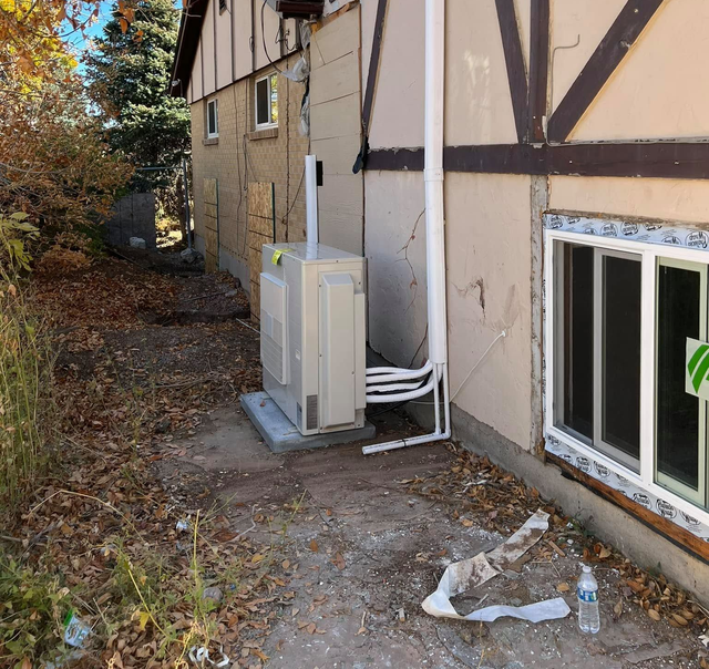 Outdoor side of a building with utility units and pipes beside a window, with fallen leaves on the ground