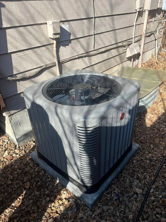 Outdoor air conditioning unit beside a gray house wall, with gravel and fallen leaves around it