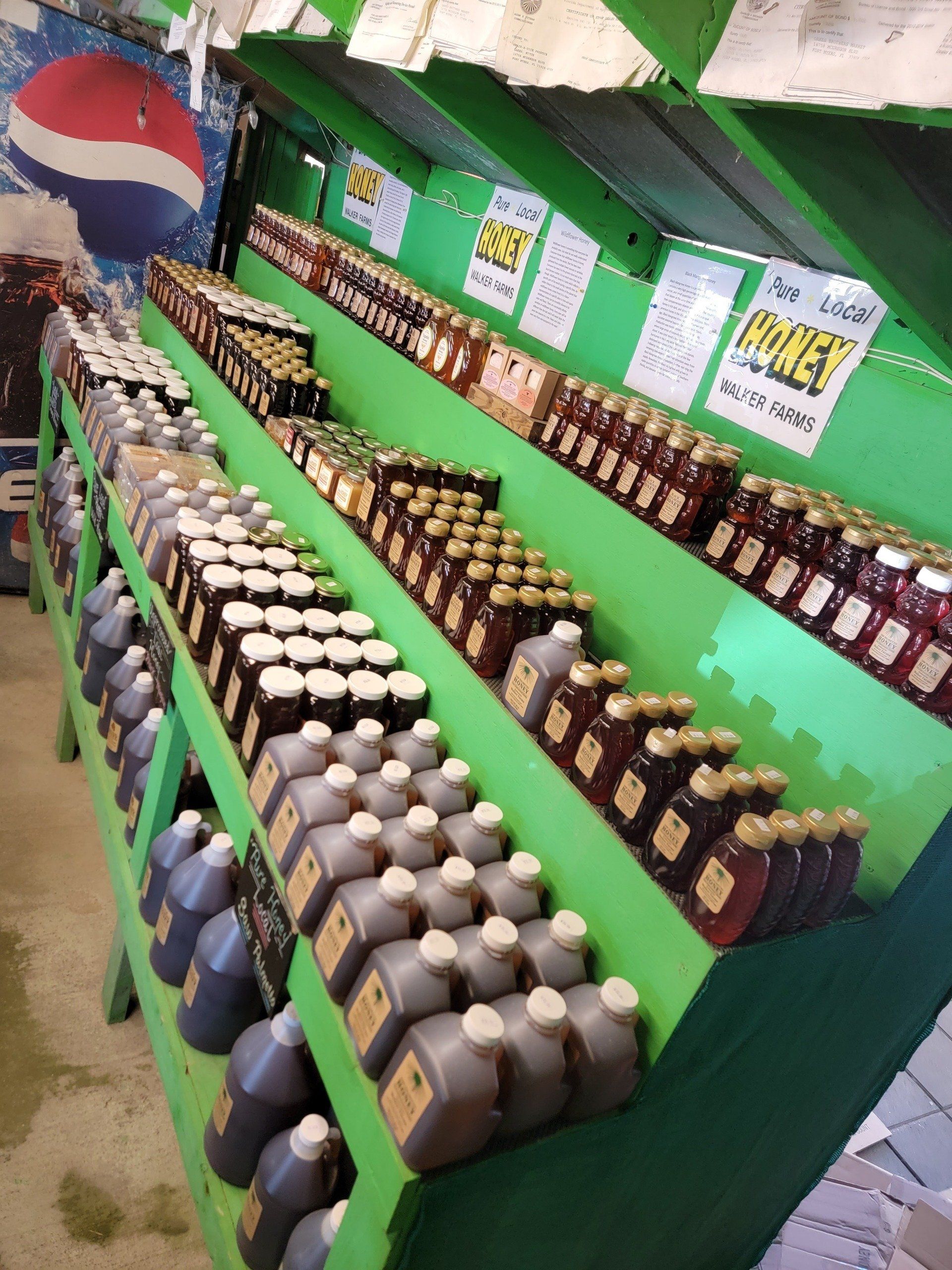 Shelves of various honey jars and jugs at a farm stand. Green shelves, Pepsi sign in background.