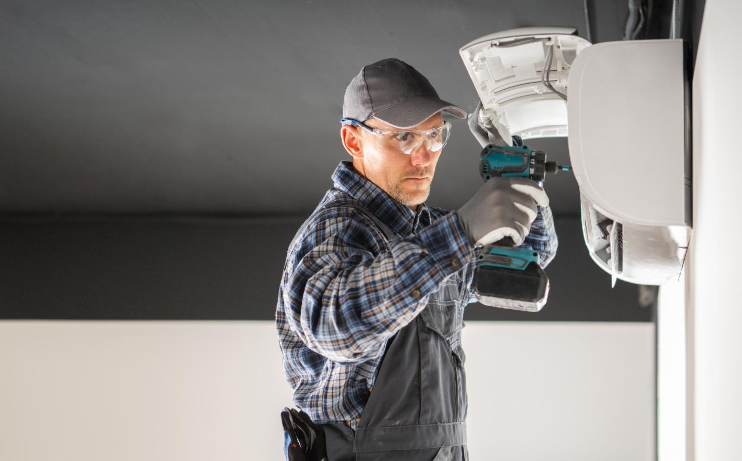 Man in work clothes uses a power drill on a white appliance attached to a wall.