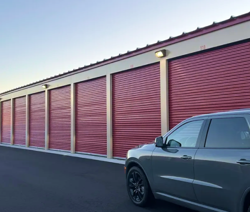A parked grey SUV in front of a row of storage units with red doors at dusk.