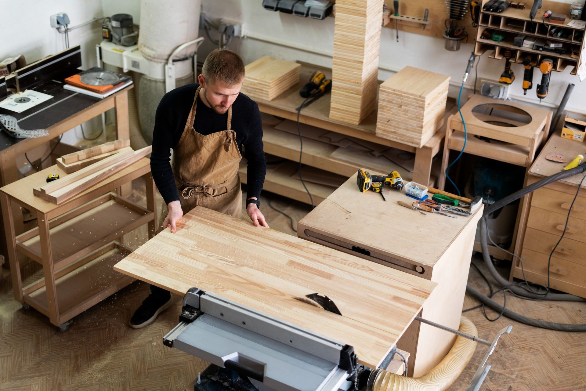 Person in an apron measuring wood in a cluttered workshop with tools and lumber.
