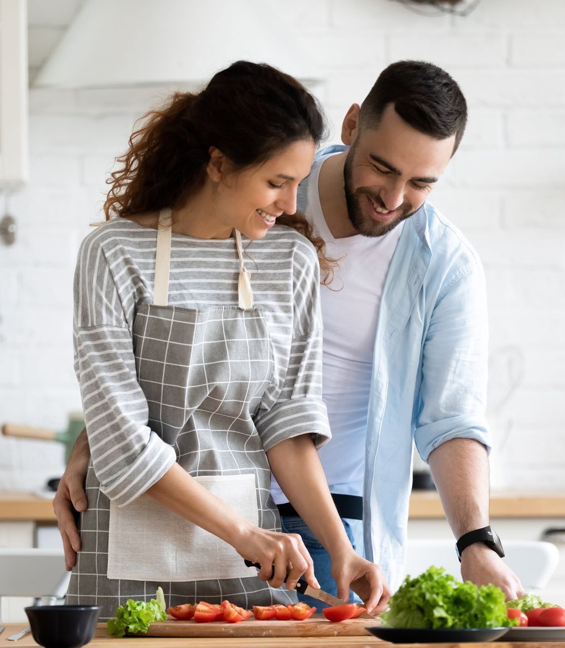 The couple stand in the kitchen cooking together, they are smiling and happy because they know the importance of date night