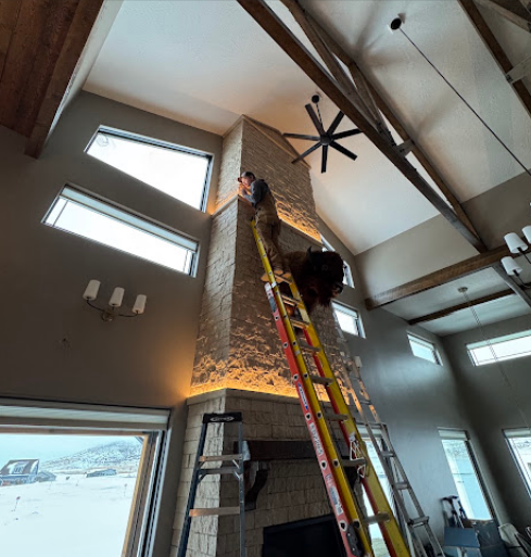 Worker on a ladder painting a high interior wall in a vaulted room with exposed beams.