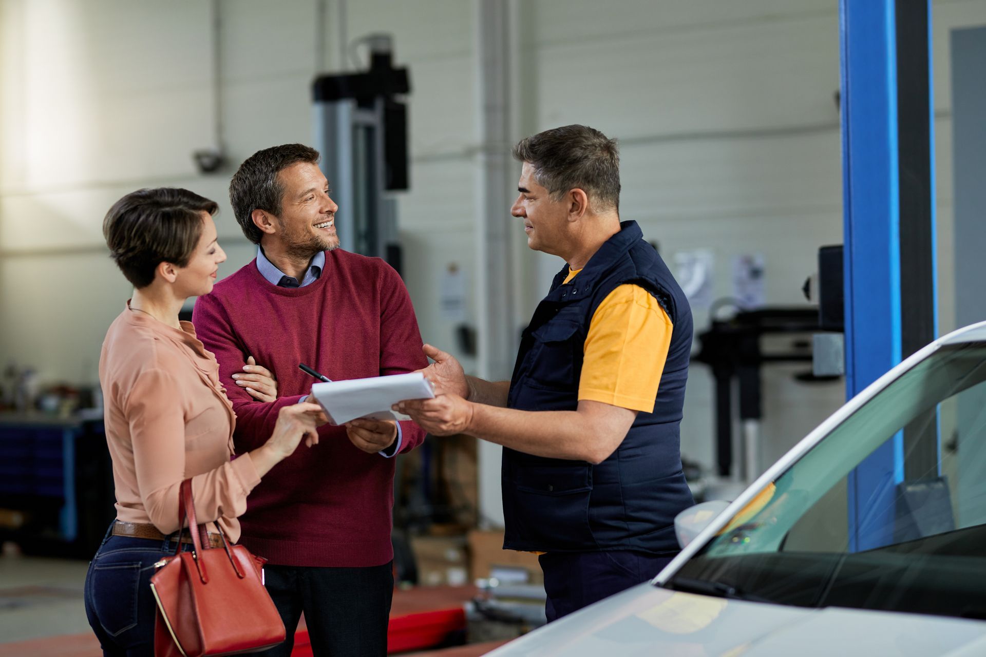 Couple talking to mechanic about car repairs in a garage. Mechanic holds paperwork; all smile.