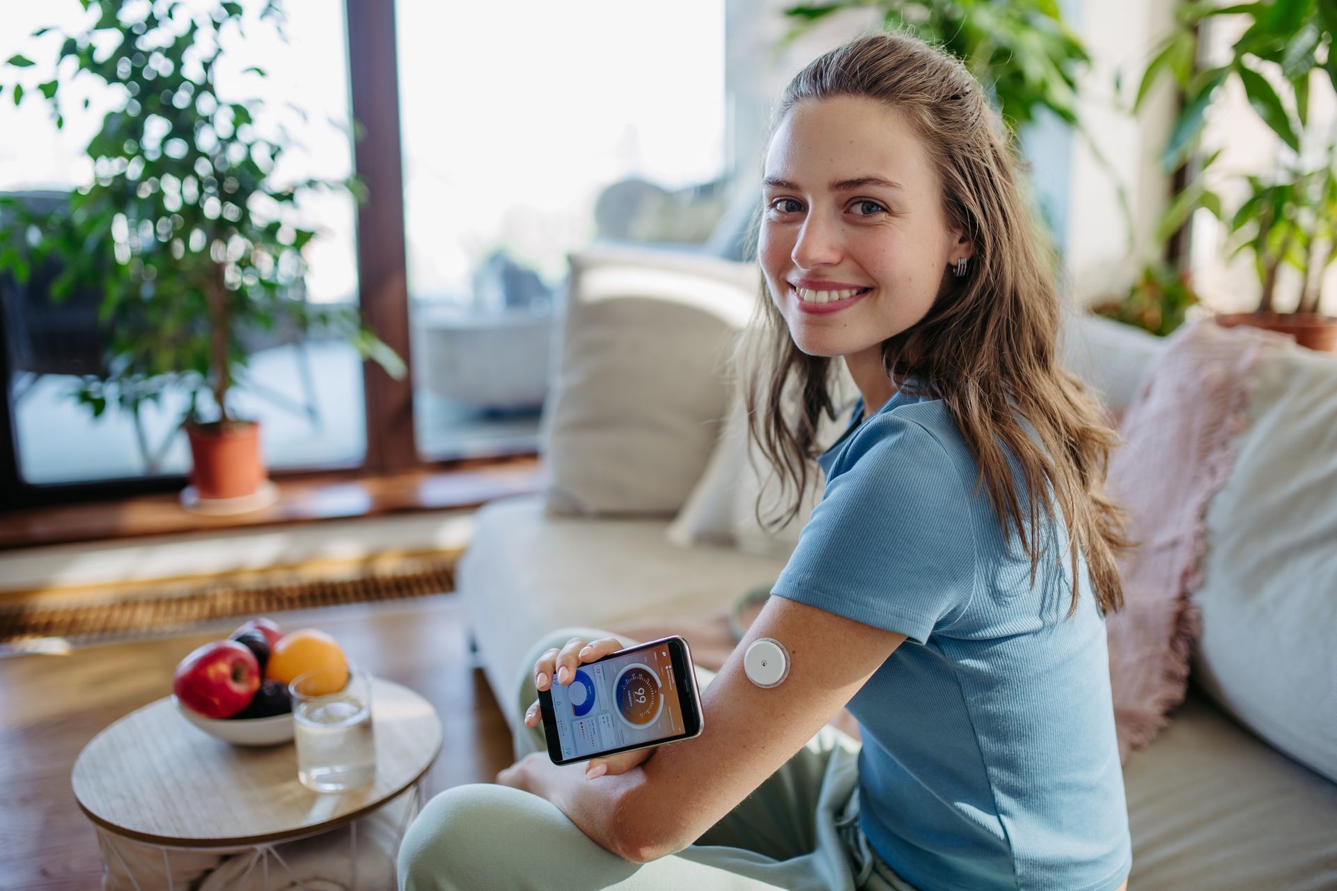 Eine Frau sitzt auf einem Sofa, hält ein Mobiltelefon in der Hand und trägt einen Sensor am Arm.