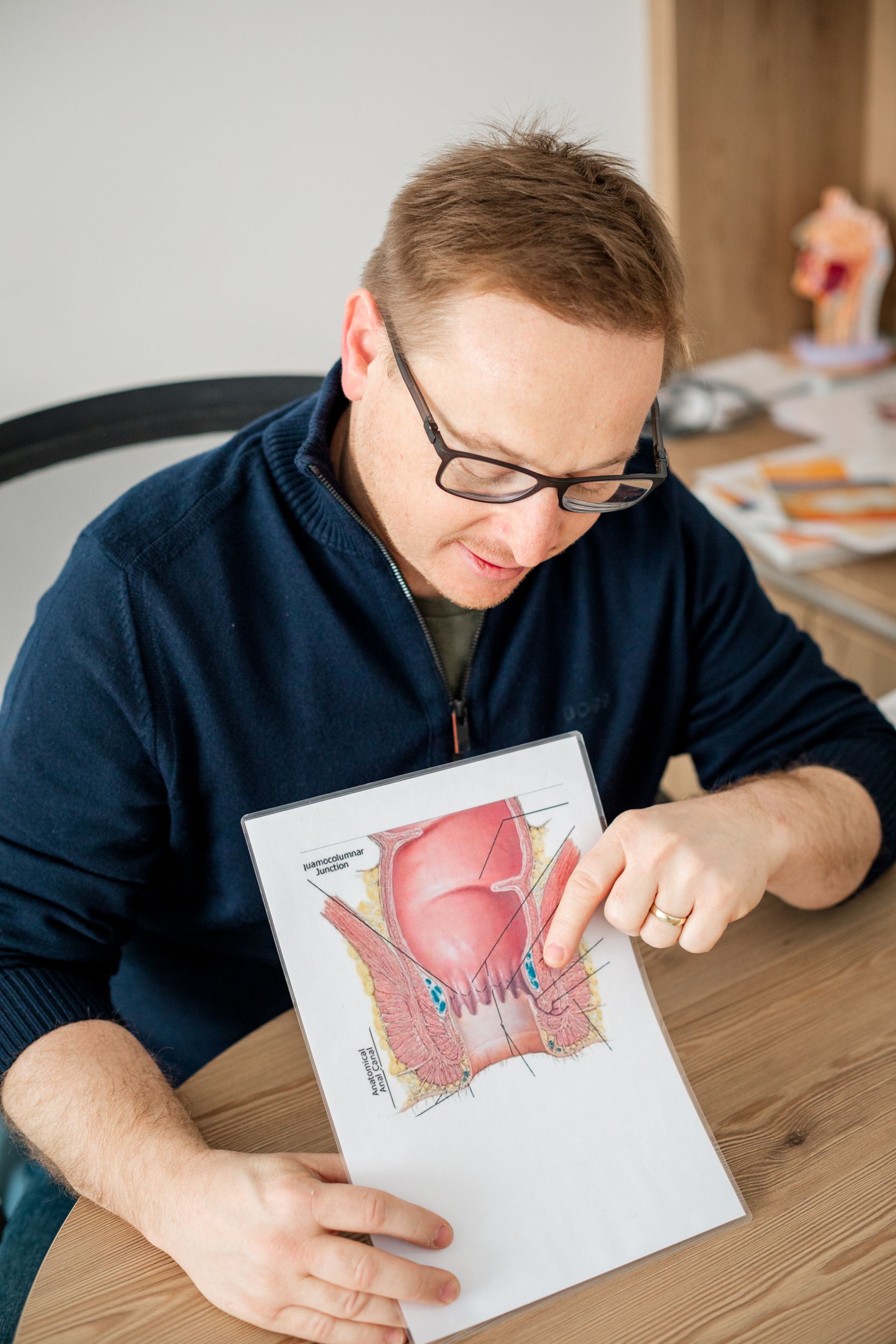 Ein Mann sitzt an einem Tisch und hält ein Blatt Papier mit einer Abbildung des männlichen Fortpflanzungssystems in der Hand.