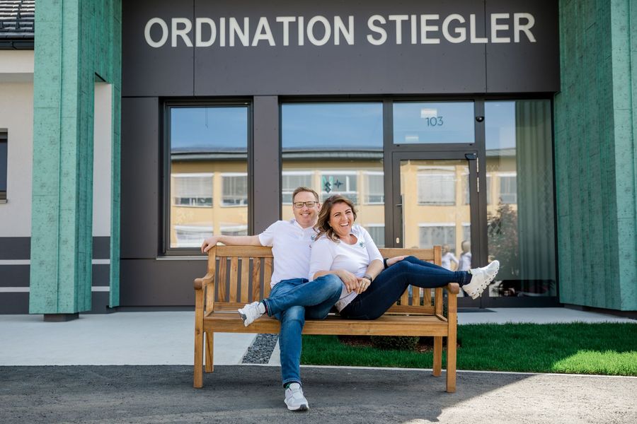 Das Team sitzt auf einer Bank vor einem Gebäude mit der Aufschrift „Ordination Stiegler“