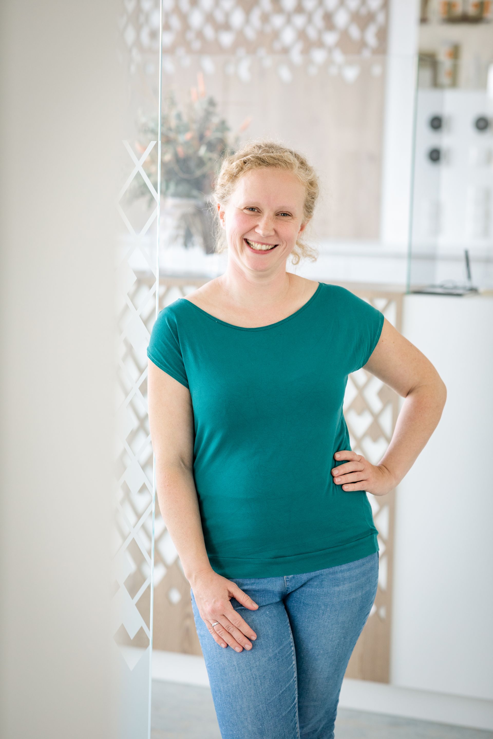 Eine Frau in einem grünen Hemd und blauen Jeans steht mit den Händen in den Hüften in einem Raum.
