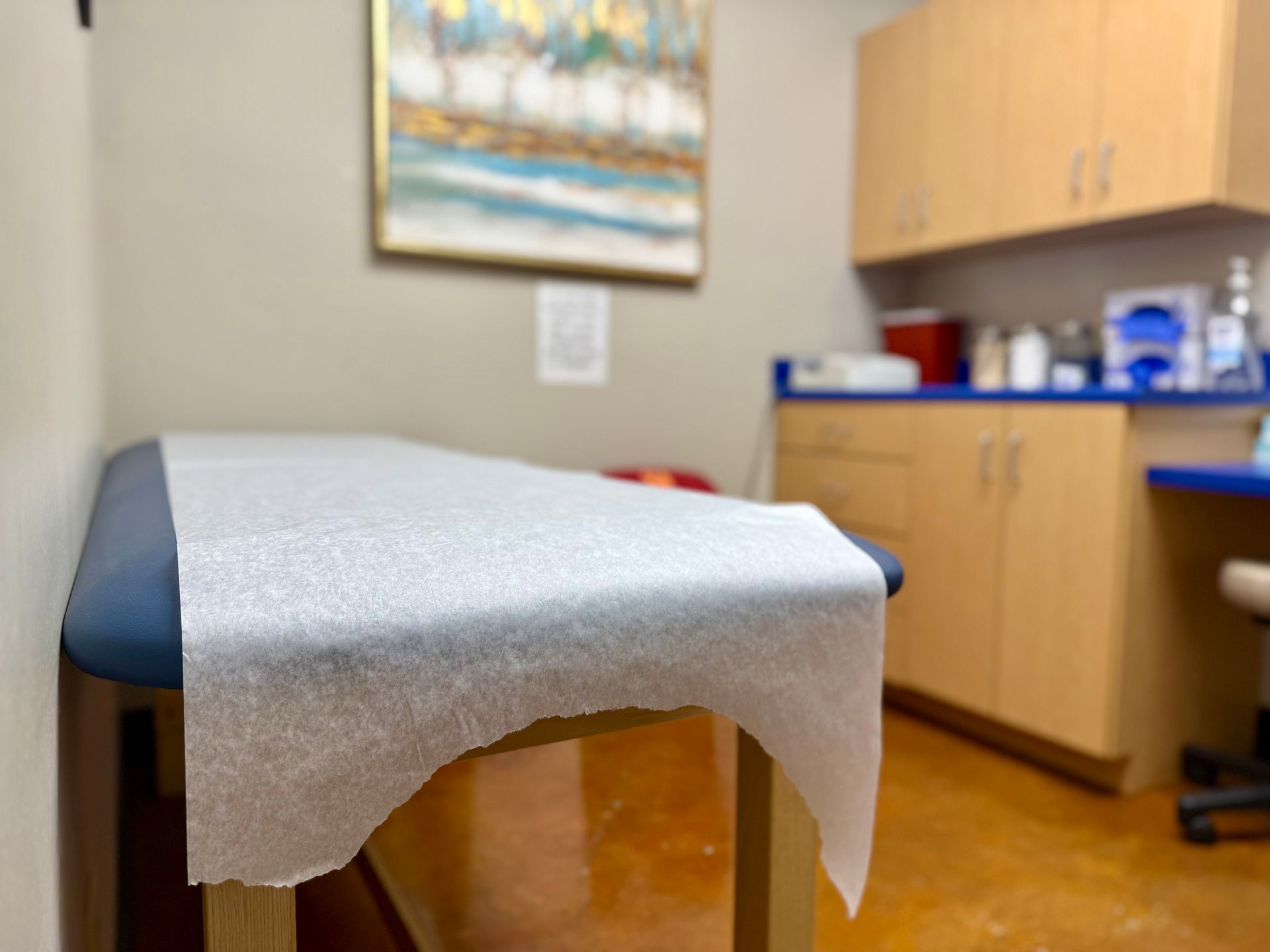 A clean, empty medical examination room featuring a treatment table covered with fresh paper, cabinets, and wall art.