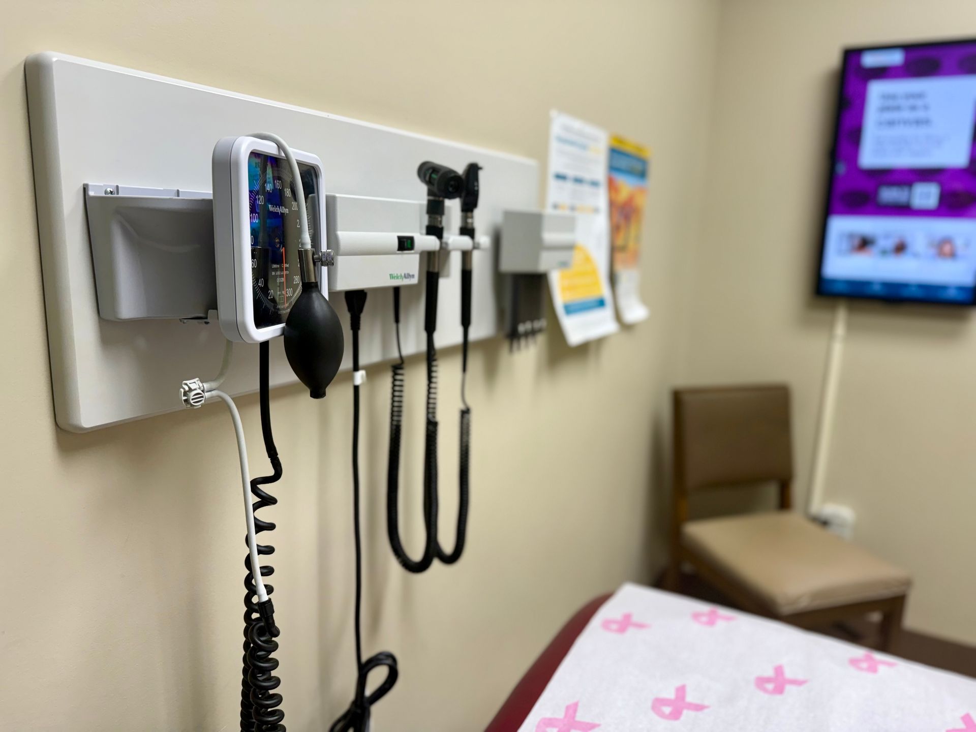 Medical diagnostic equipment mounted on a clinic wall next to an examination table with a pink ribbon patterned cover.