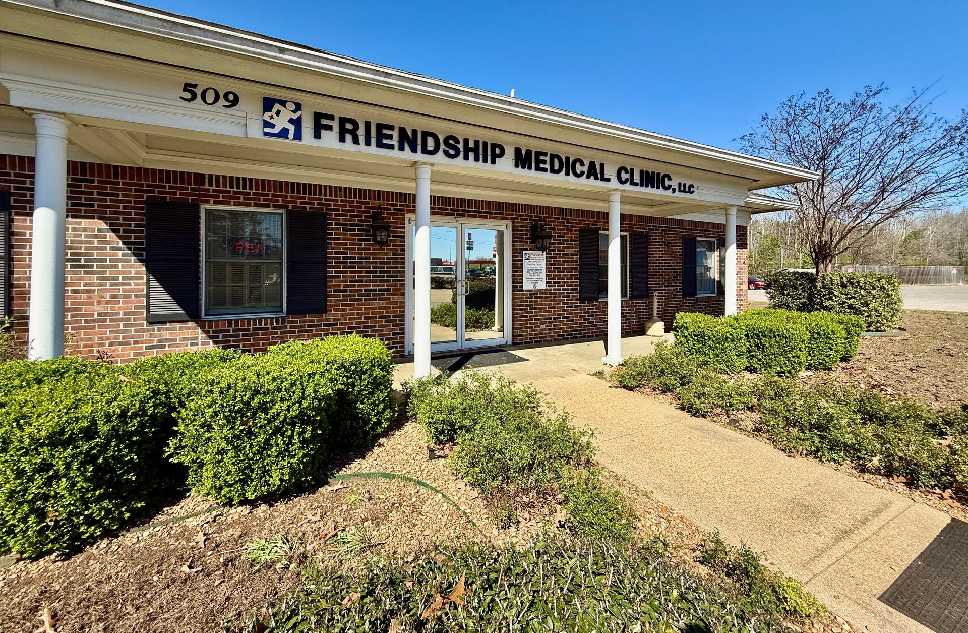 Friendship Medical Clinic building with a brick exterior, white columns, and a paved walkway, under a clear blue sky.