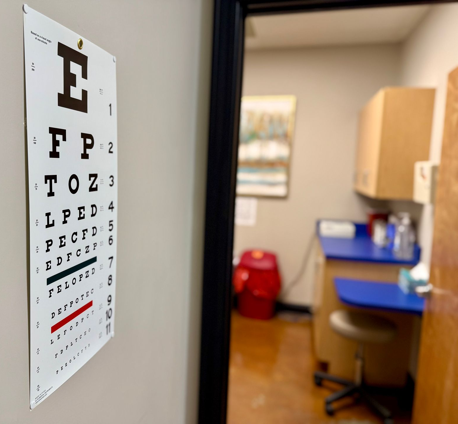 A Snellen eye chart hangs on a wall next to a doorway looking into an exam room with a counter, stool, and medical supplies.