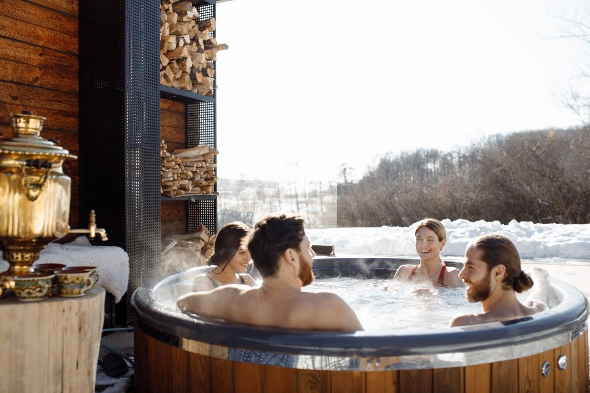 People relaxing in a steaming outdoor hot tub beside a brick lodge, overlooking a snowy landscape