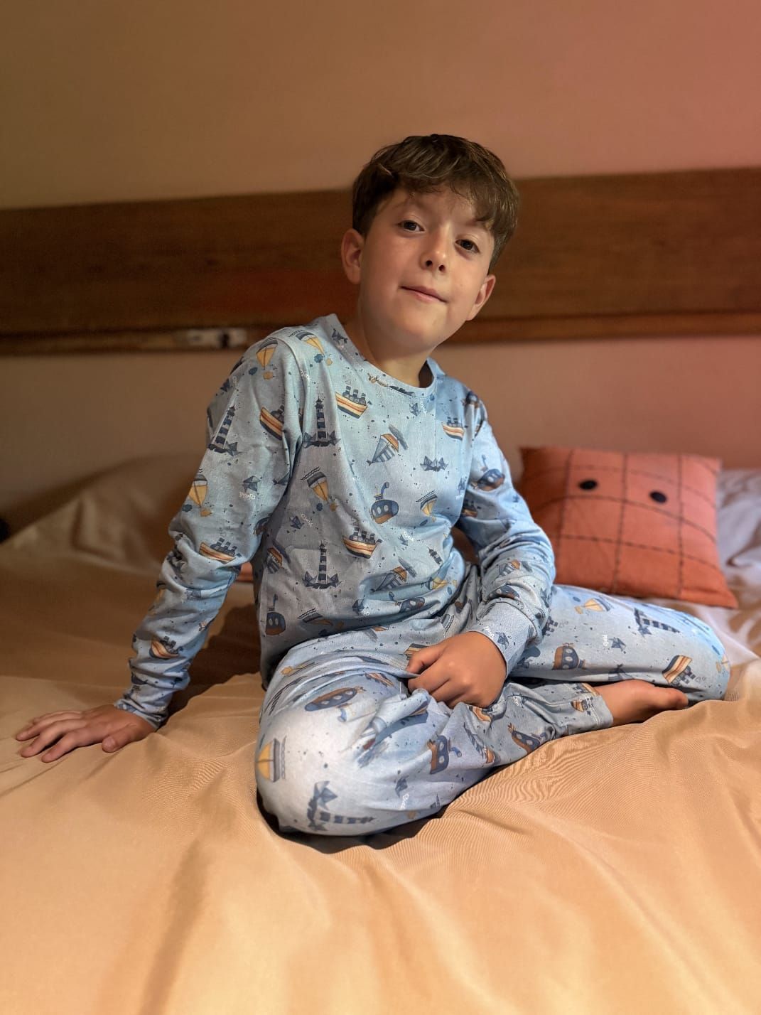 Niño pequeño sonriendo, vistiendo un pijama azul de temática náutica, de pie afuera en un patio.