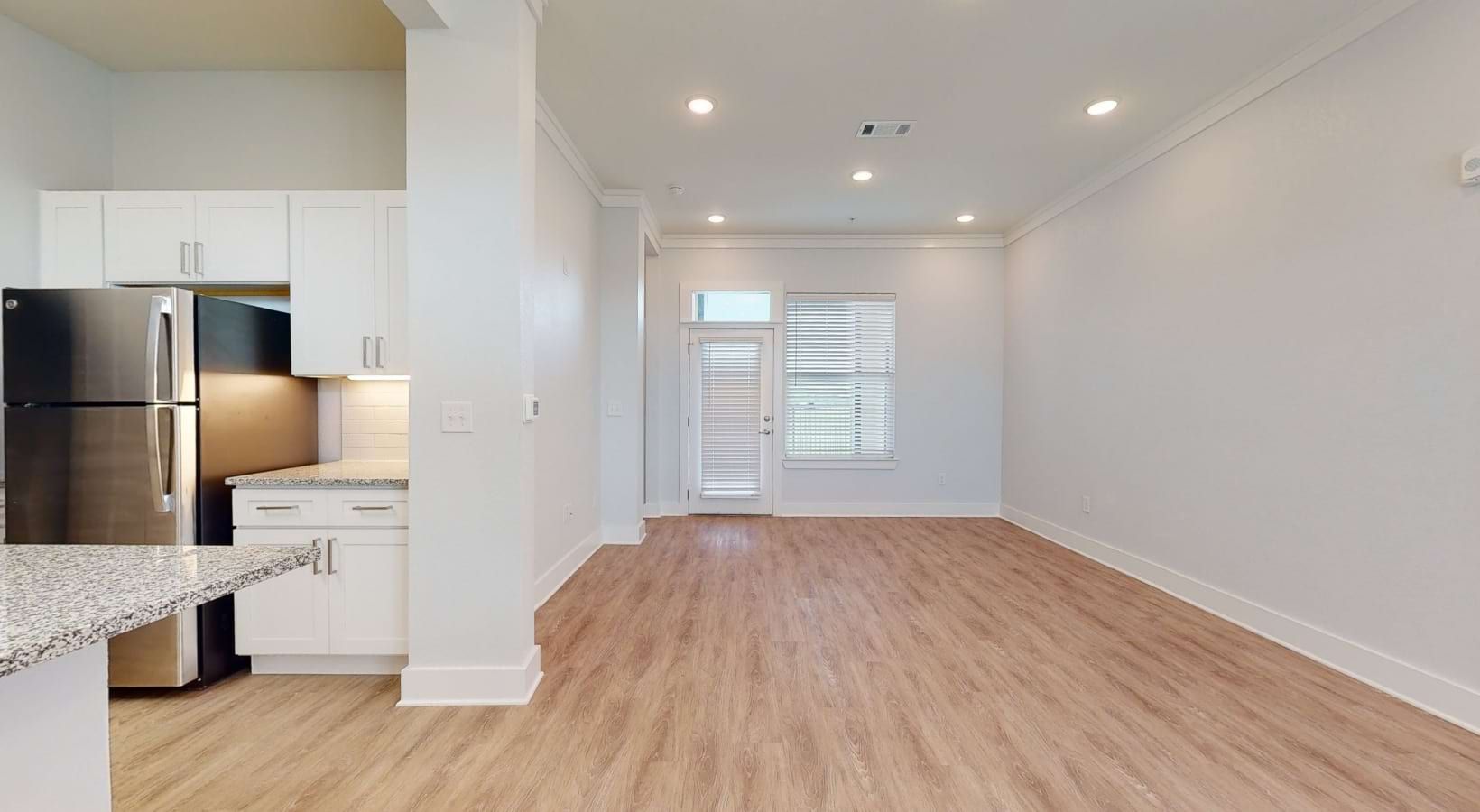 Bright, empty room with a kitchen, white walls, and wood-look flooring. A stainless steel refrigerator is visible.