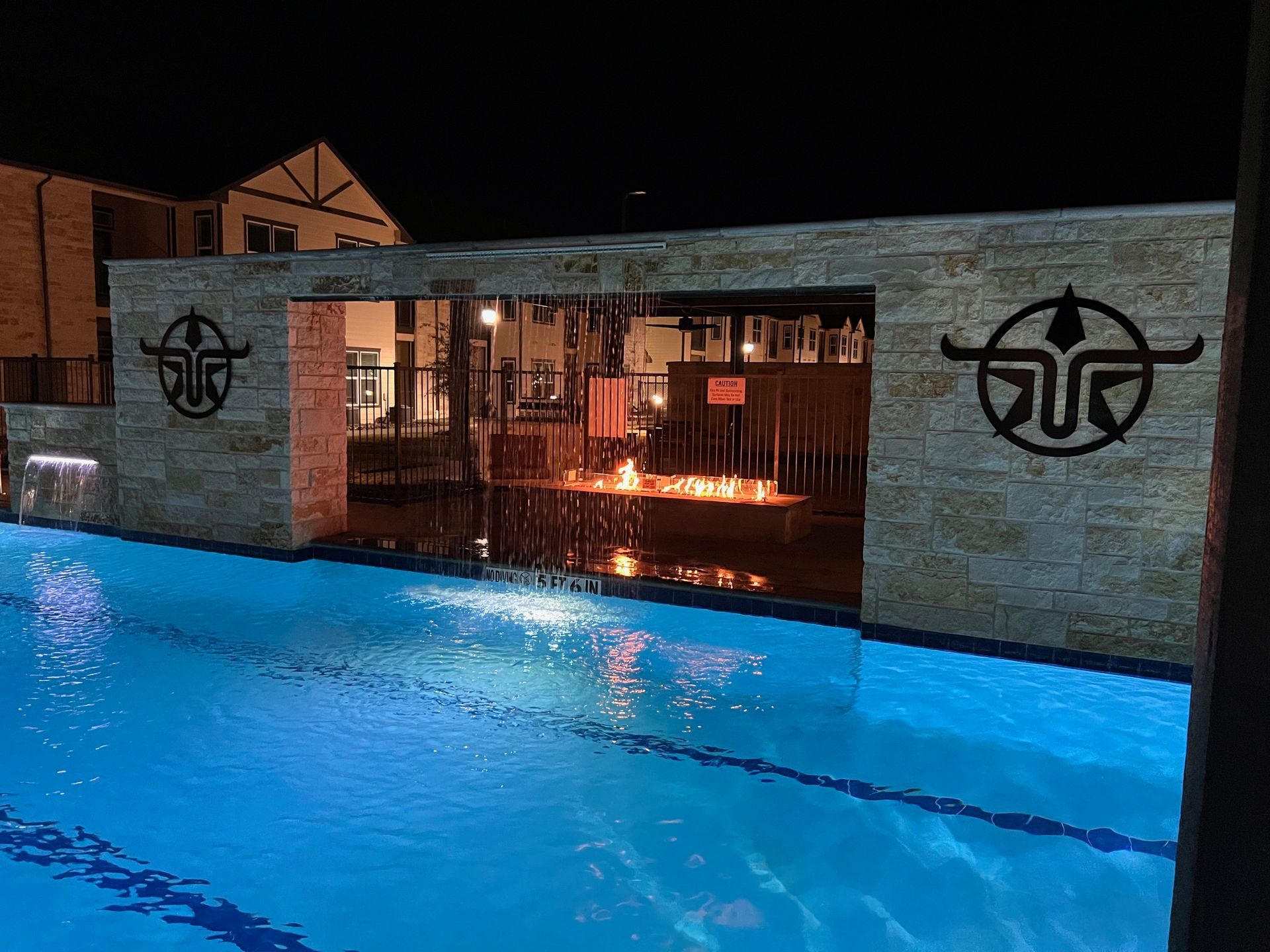 A night view of a pool with a water feature and lit wall in front of a building.