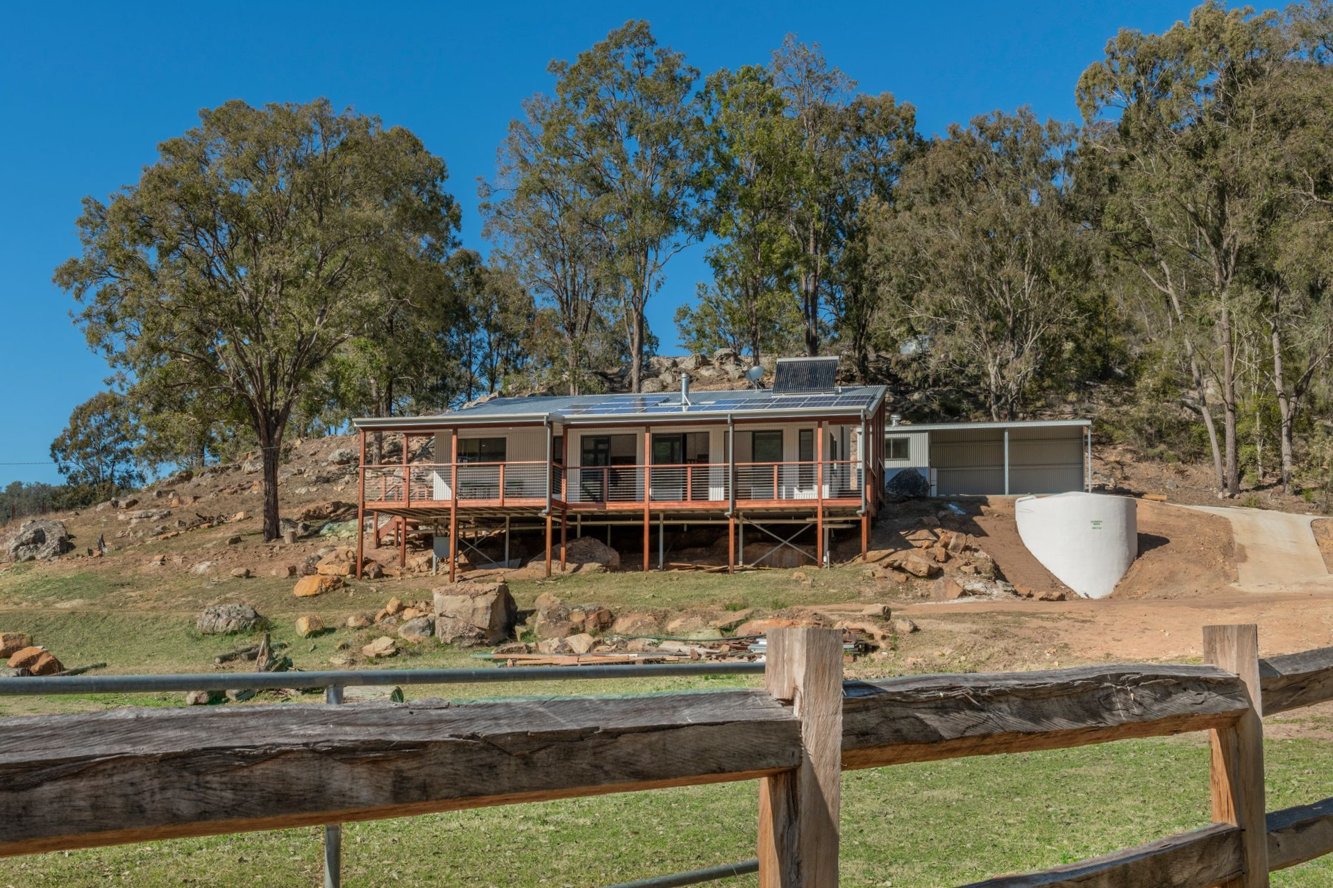 Cottage on a hillside with a wooden deck, surrounded by trees and a fence — Indie Living In Hunter Valley, NSW