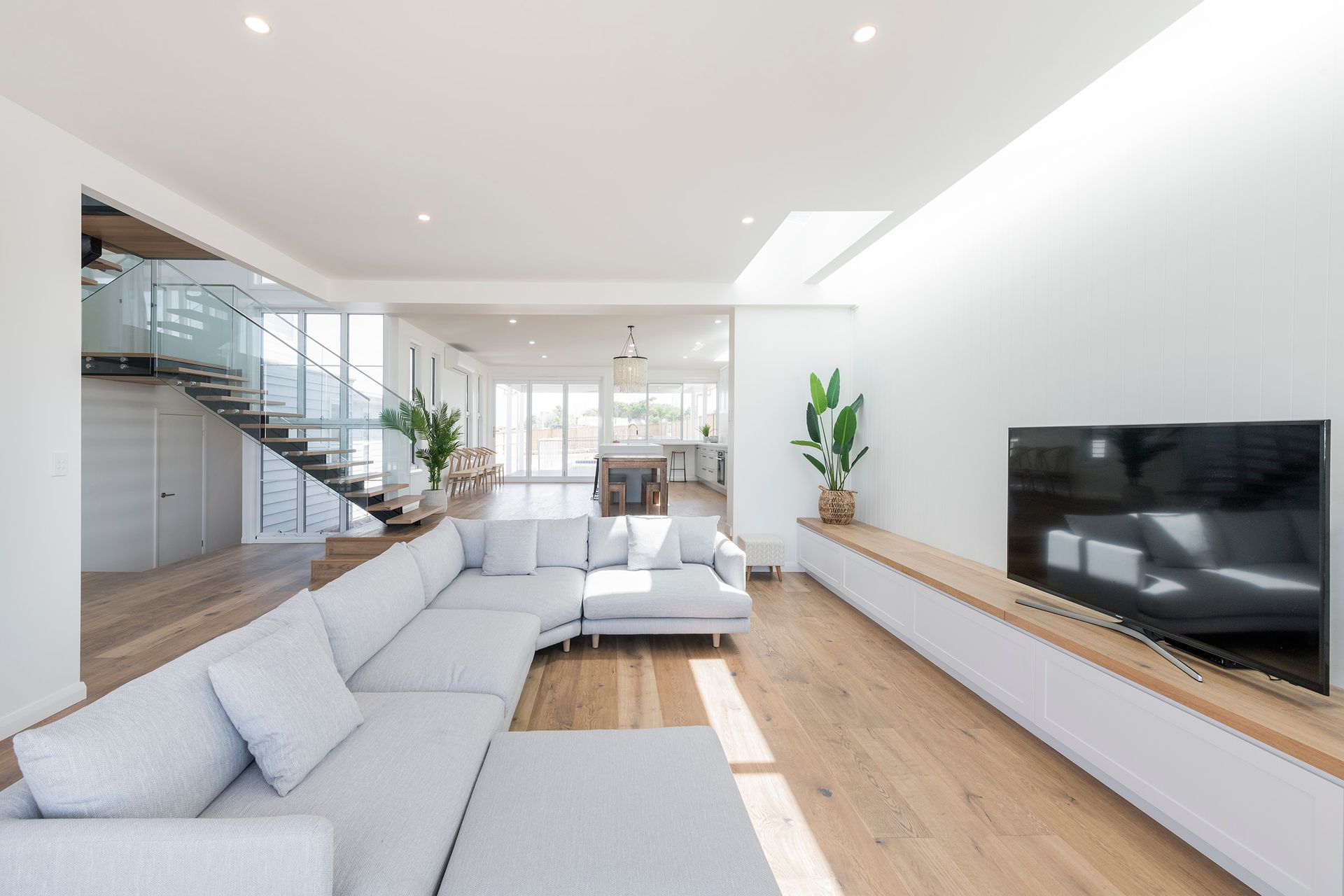 Modern living room with light-colored sofa, wood floors, white walls, TV on a built-in cabinet, and a staircase — Indie Living In Bennetts Green, NSW