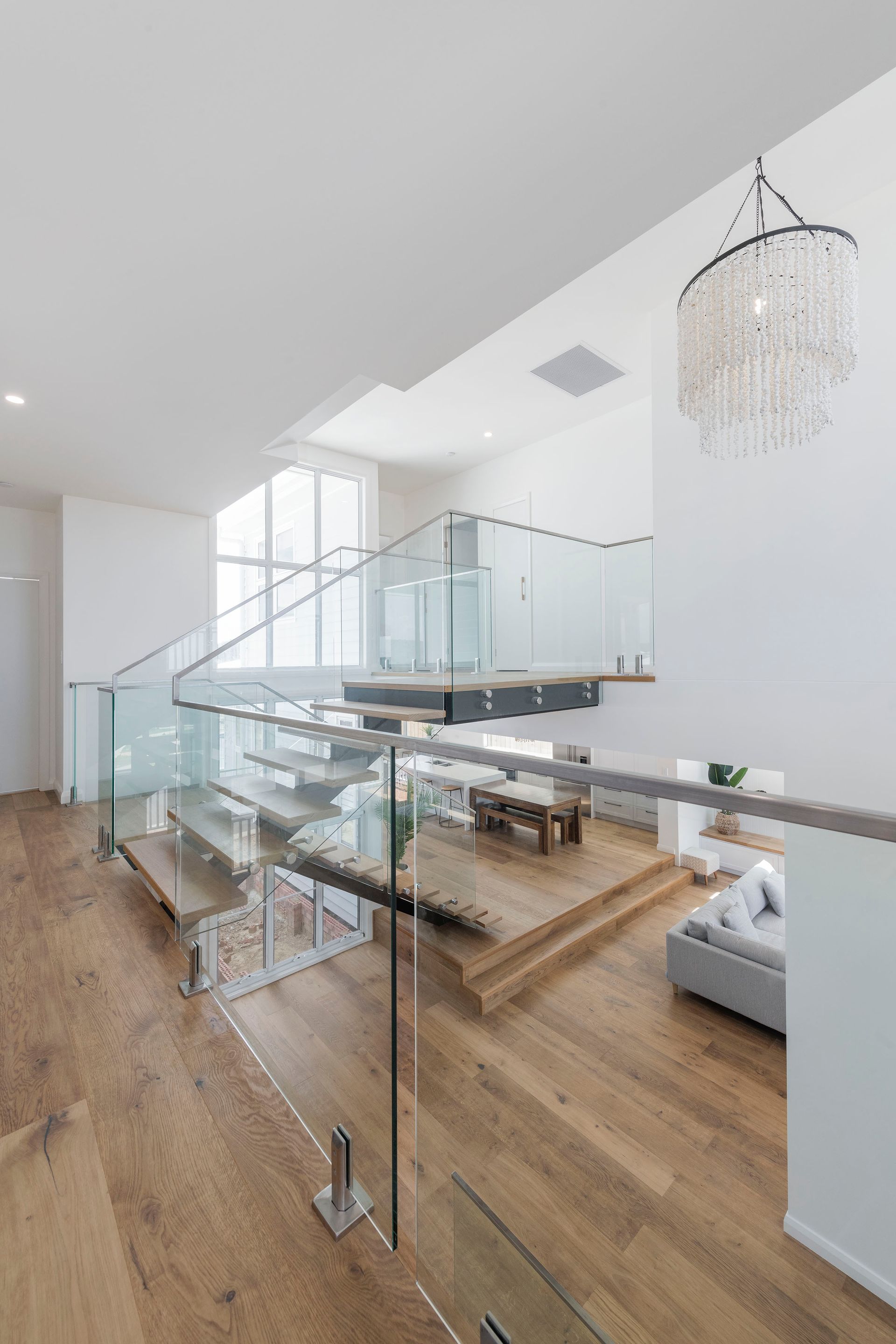 Modern, two-story interior with glass-railed stairs, wood floors, and a chandelier — Indie Living In Bennetts Green, NSW
