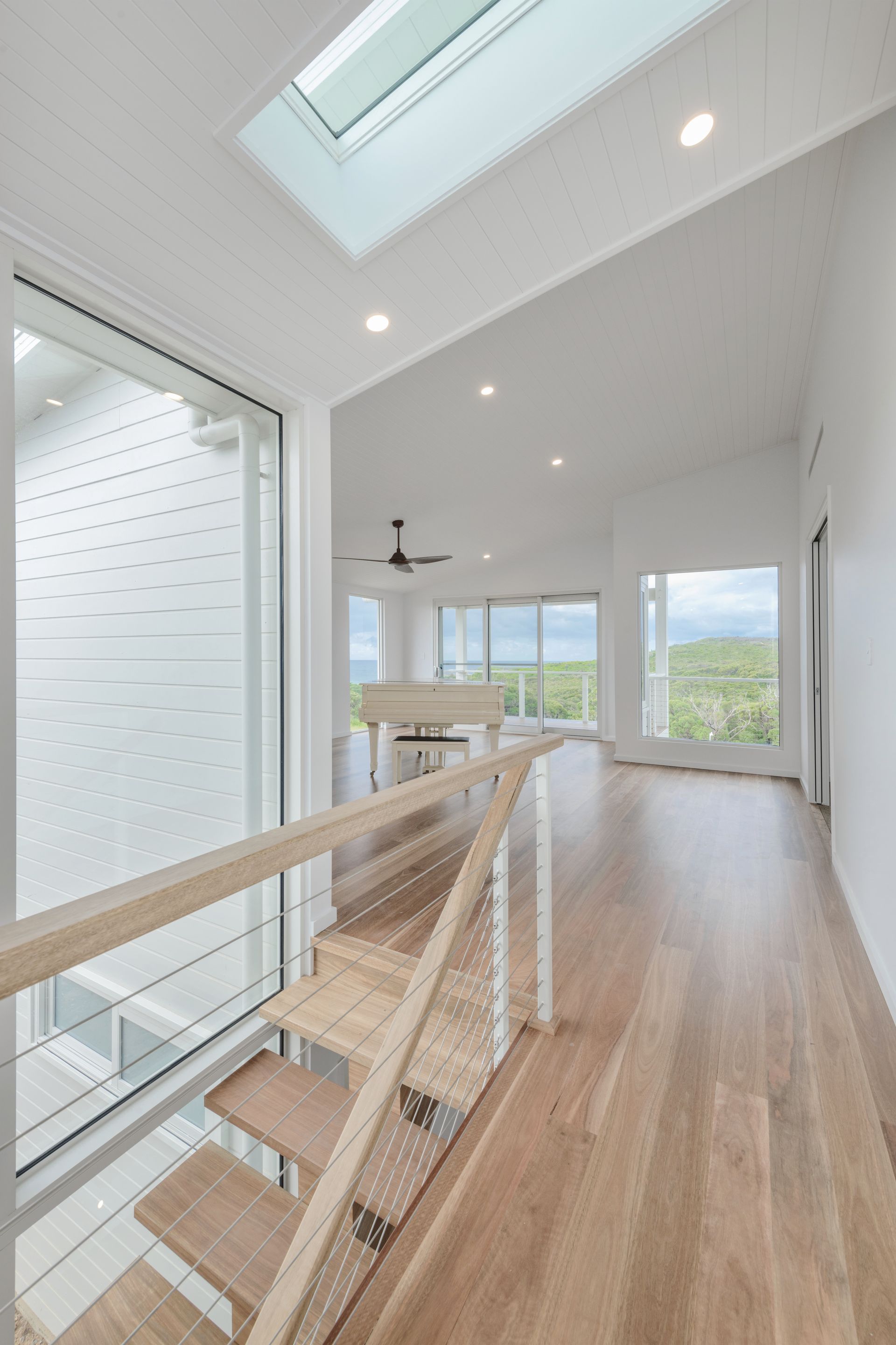 Bright, modern hallway with wooden stairs, skylight, and view of ocean through windows — Indie Living In Bennetts Green, NSW