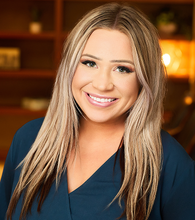 Woman with long blonde hair, smiling, wearing a dark blue top, interior setting.